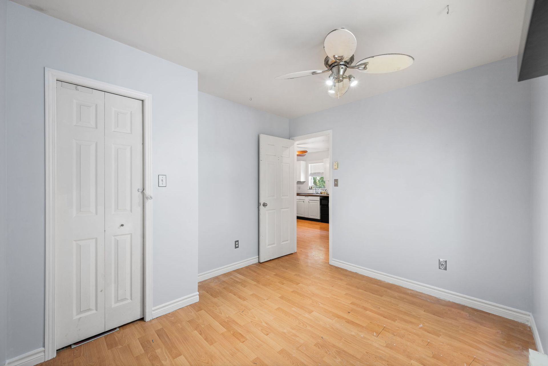 Empty room with pale blue walls, white doors, and wood flooring; a doorway leads to a kitchen.