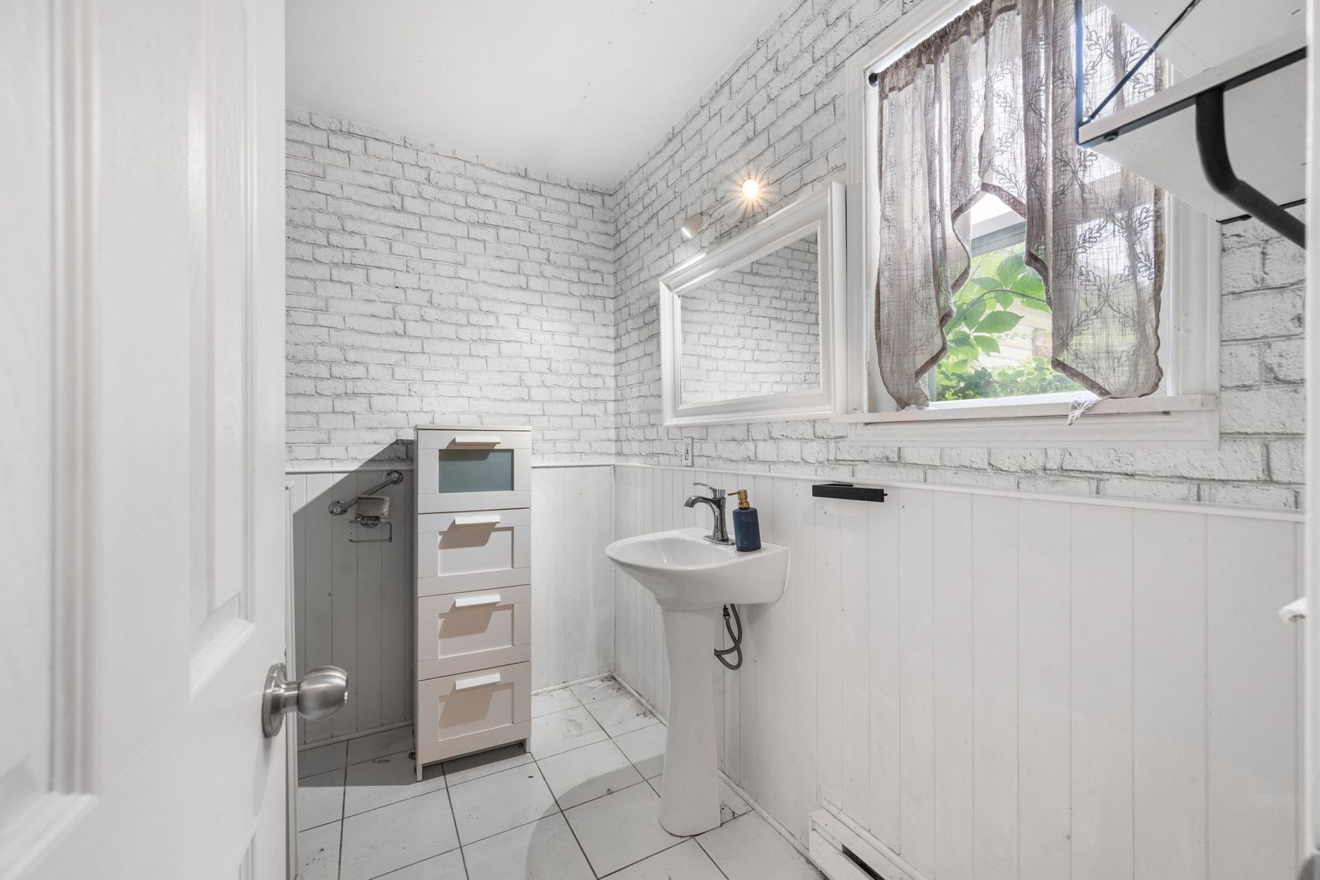 White brick-walled bathroom with pedestal sink, a window with sheer curtain, and storage cabinet.