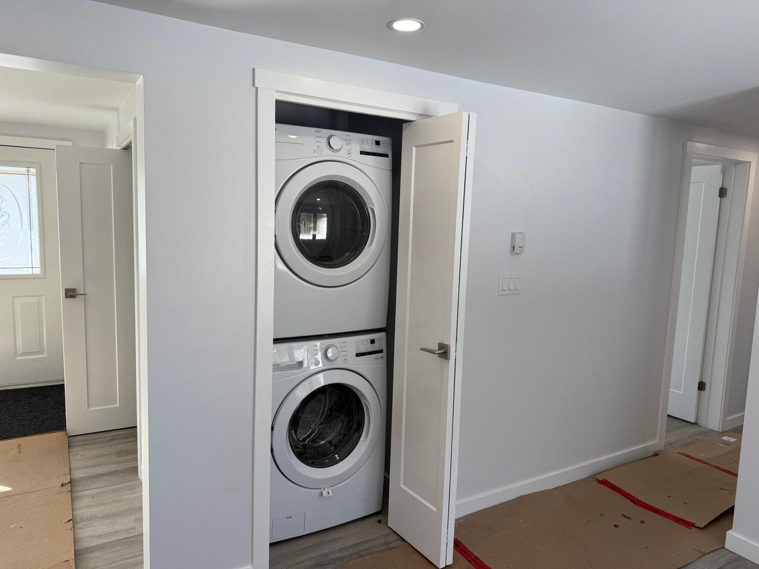 Stacked white washer and dryer in a closet within a white-walled hallway with light wood flooring.