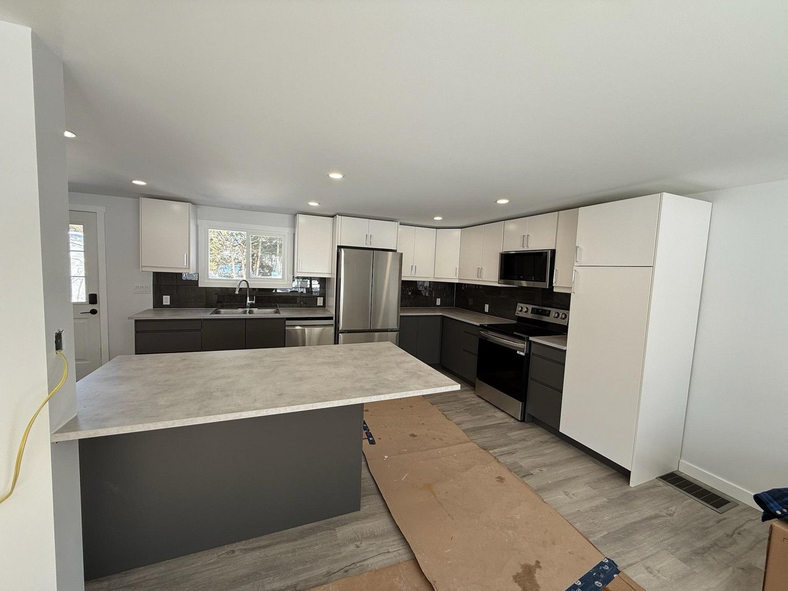 Modern kitchen with a gray island, dark lower cabinets, white upper cabinets, stainless appliances, and wood-look floors.