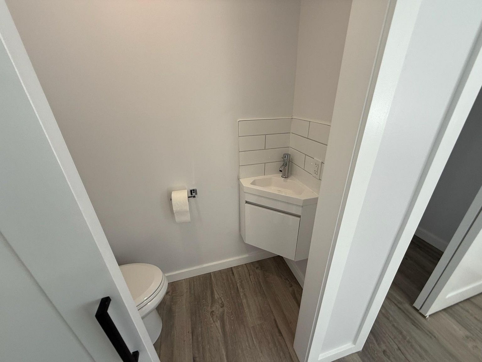 Small half-bathroom with light gray walls, wood-look flooring, white toilet, and a white corner wall-mount vanity.