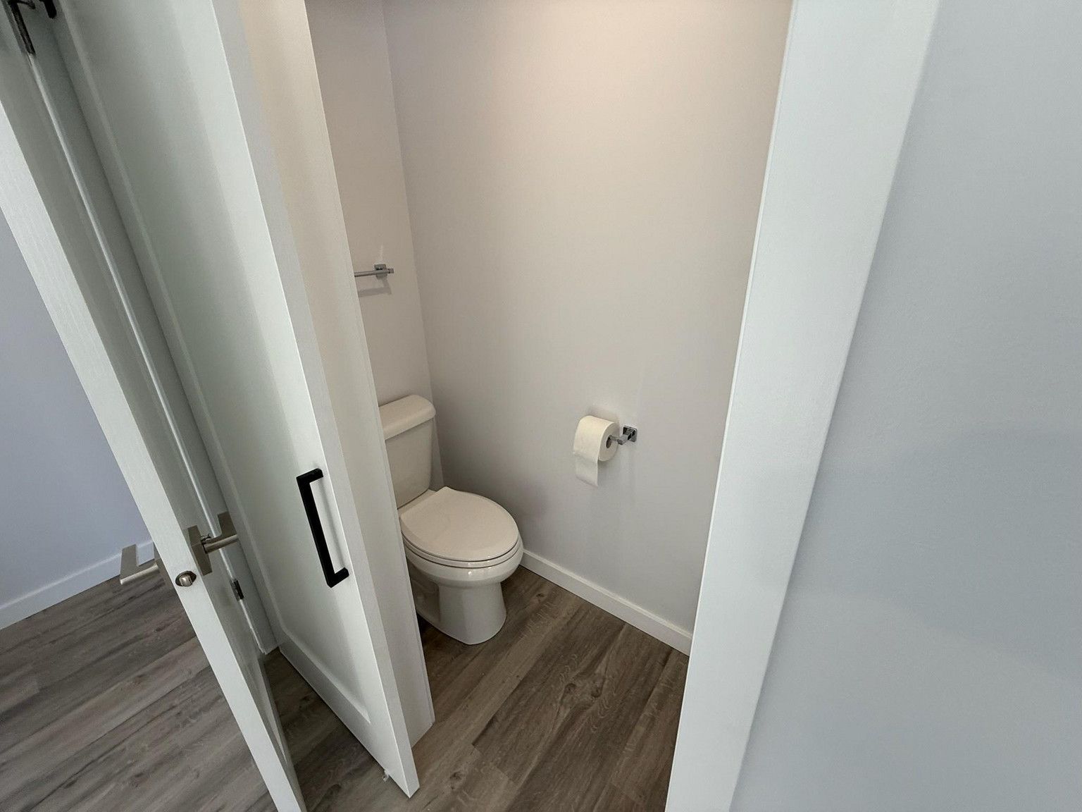 A small, white-walled bathroom with wood-grain flooring, featuring a toilet and a wall-mounted toilet paper holder.