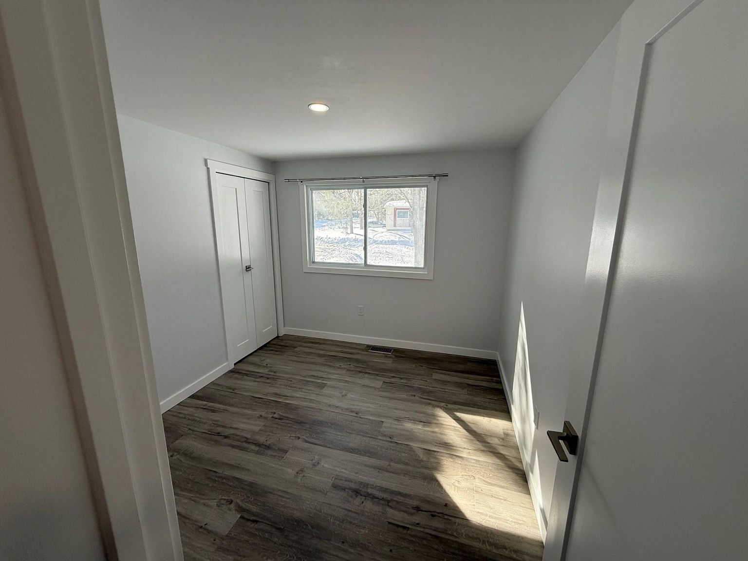 A small, empty room with light gray walls, white trim, and wood-patterned flooring, featuring a window and a closet.