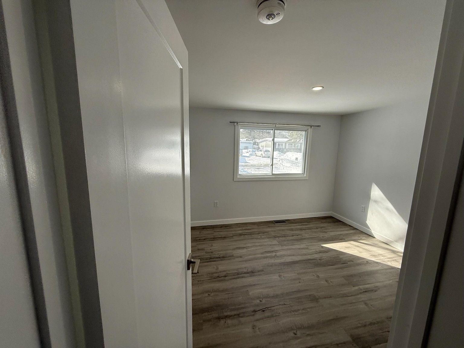 An empty bedroom with light gray walls, wood-look flooring, and a window centered on the far wall.