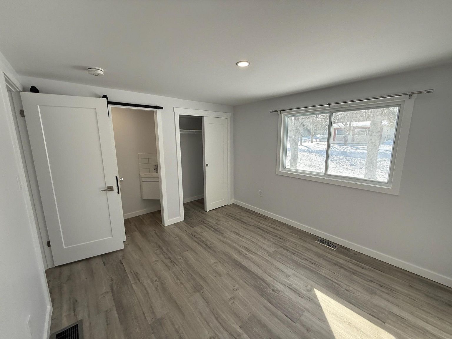 An empty bedroom with light grey walls, wood-look flooring, a closet, a bathroom doorway, and a window with snow outside.