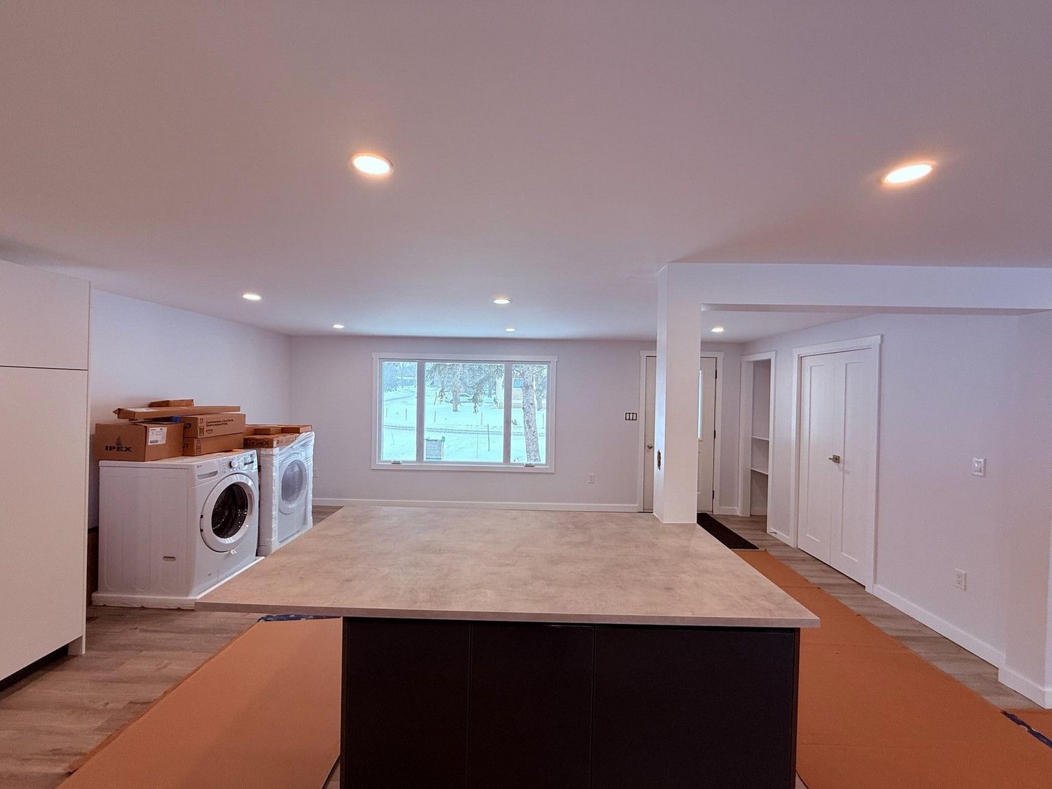 A kitchen island with a light countertop sits in a basement with light walls, recessed lights, and laundry appliances.