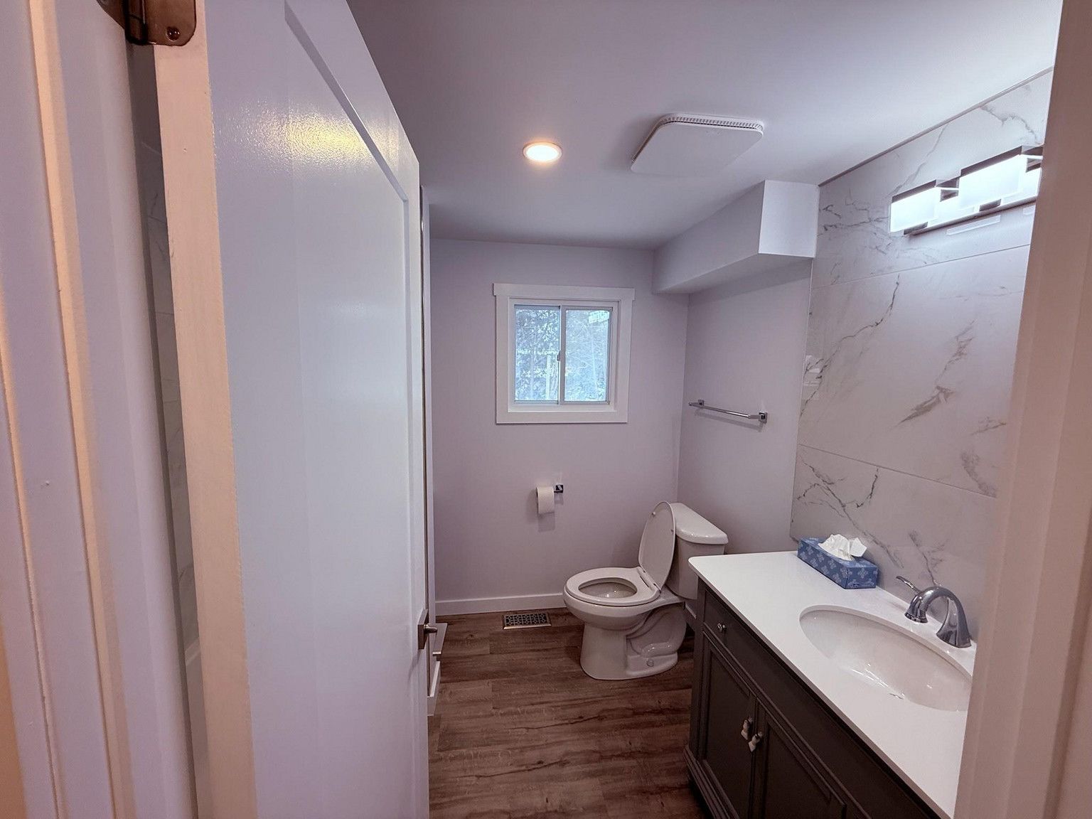 A bright, modern bathroom with grey walls, white vanity, marble-patterned backsplash, toilet, and wood-look flooring.