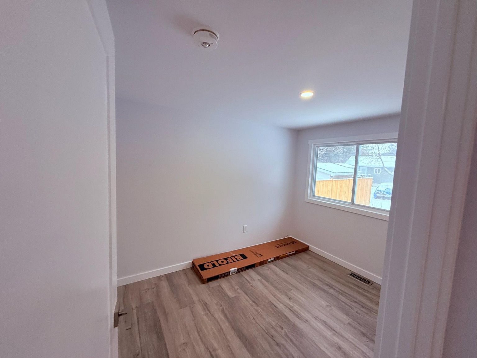 Empty bedroom with light grey walls, light wood-look flooring, a window, and a flat-packed box on the floor.
