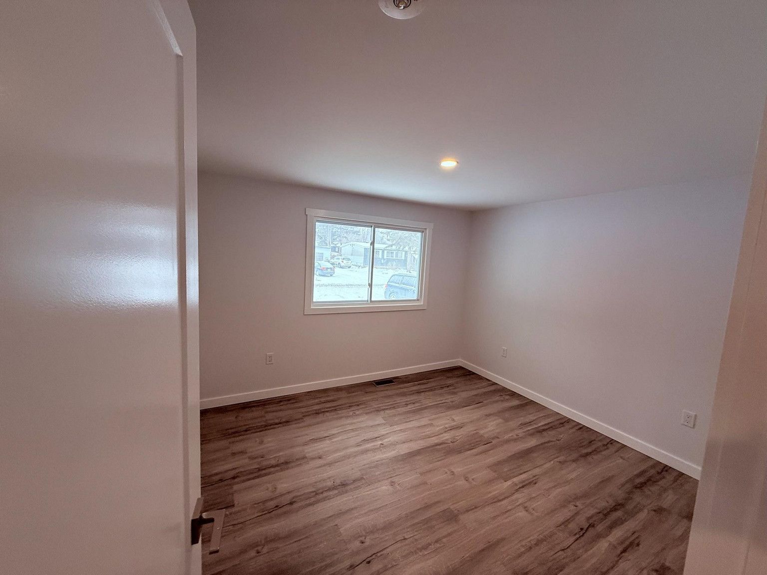 A bright, empty bedroom with light-colored walls and wood-patterned flooring, featuring a single window on the back wall.