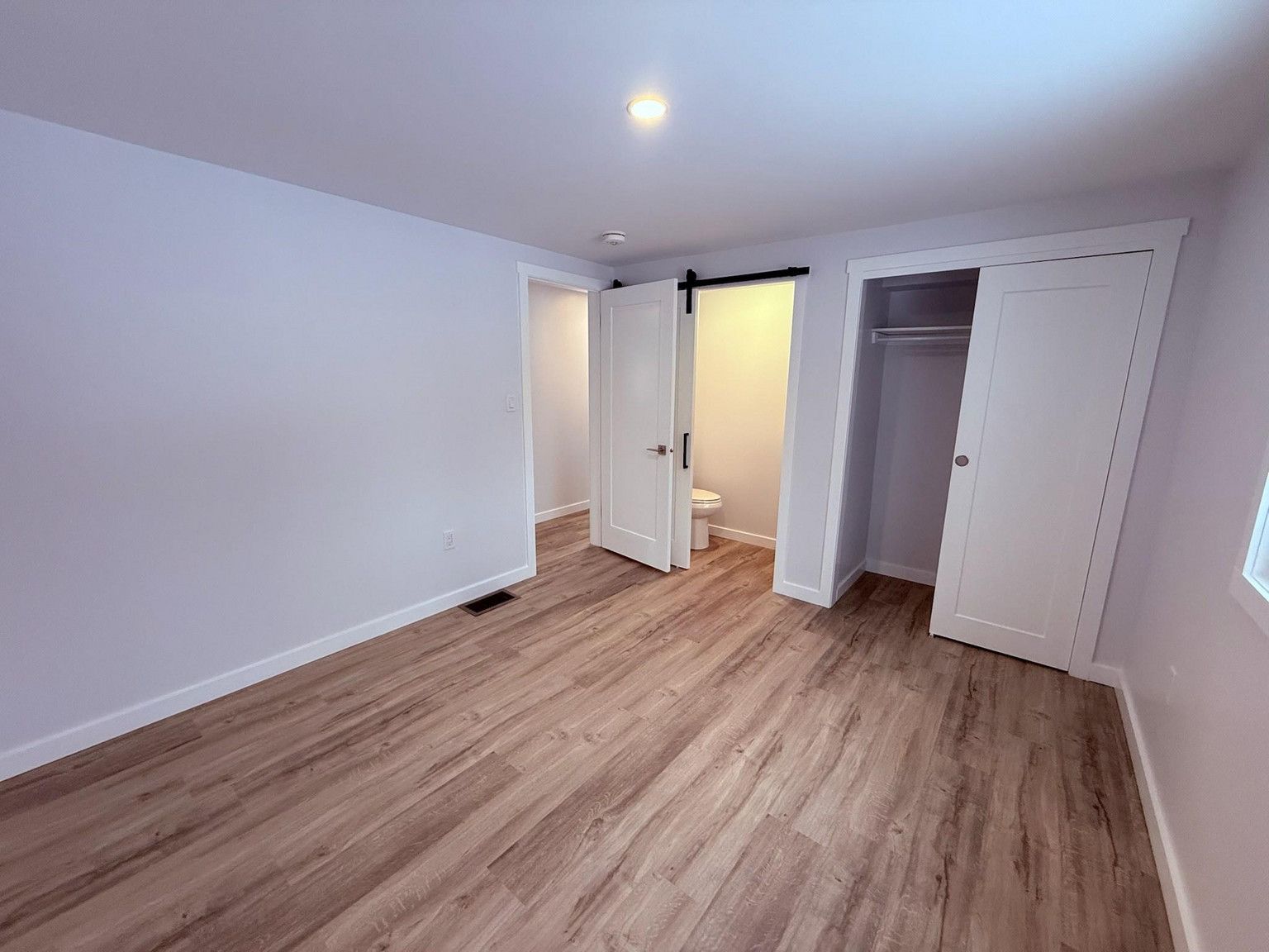 A room with light wood flooring, light grey walls, a white door leading to a bathroom, and an open closet.