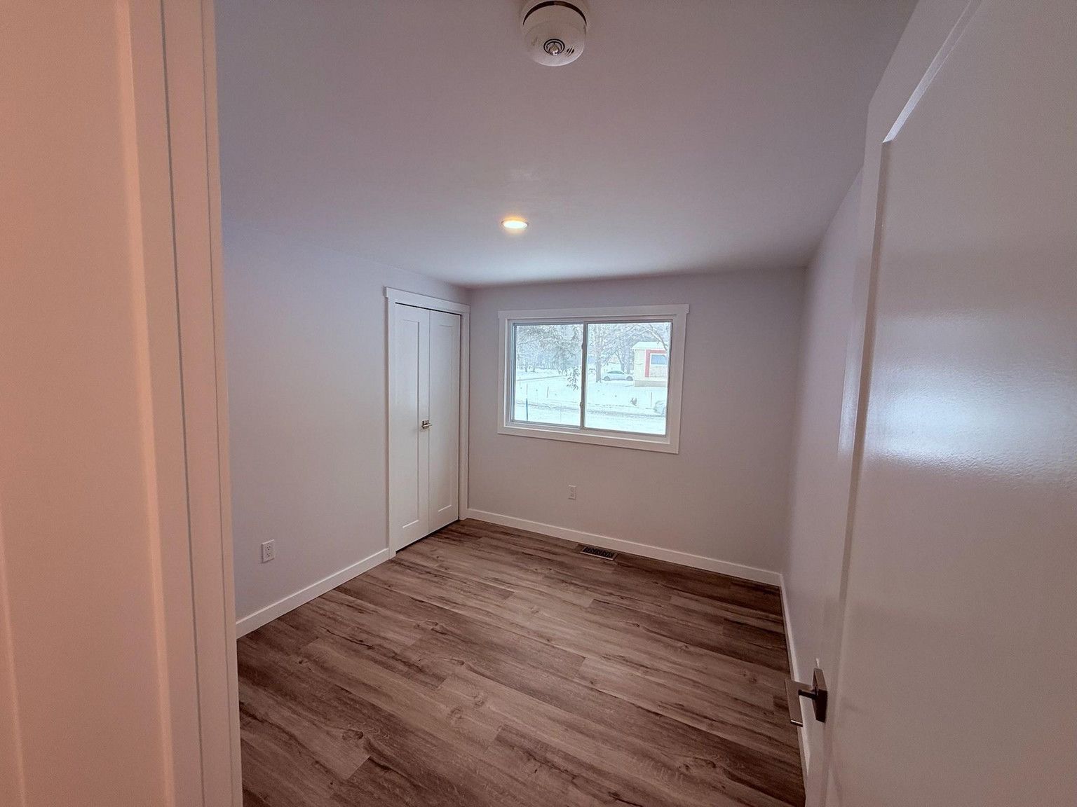 A small, empty bedroom with light grey walls, wood-look flooring, a white closet door, and one window.
