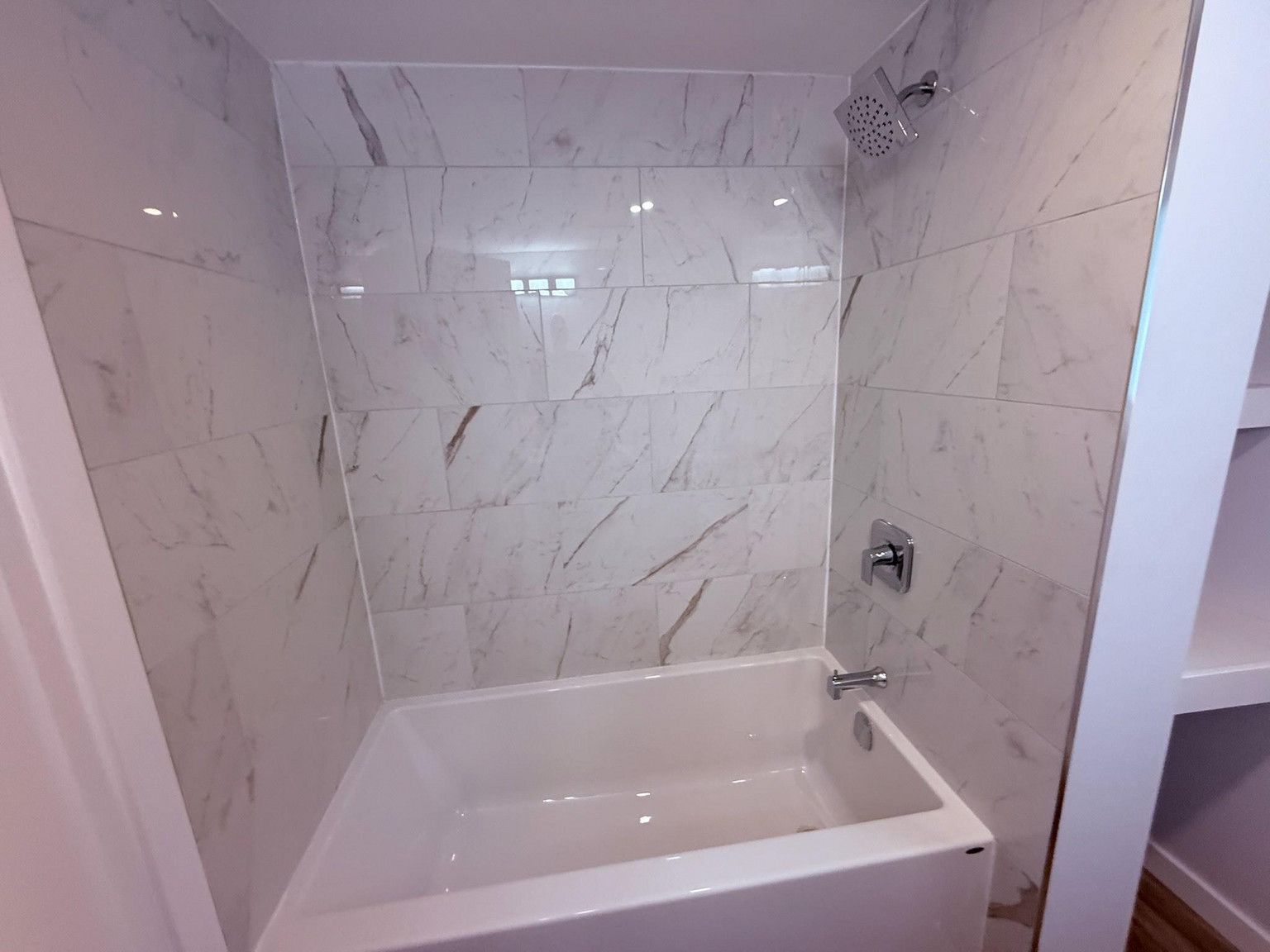 A white bathtub with a chrome shower head and faucet, surrounded by white marble-patterned tile walls.