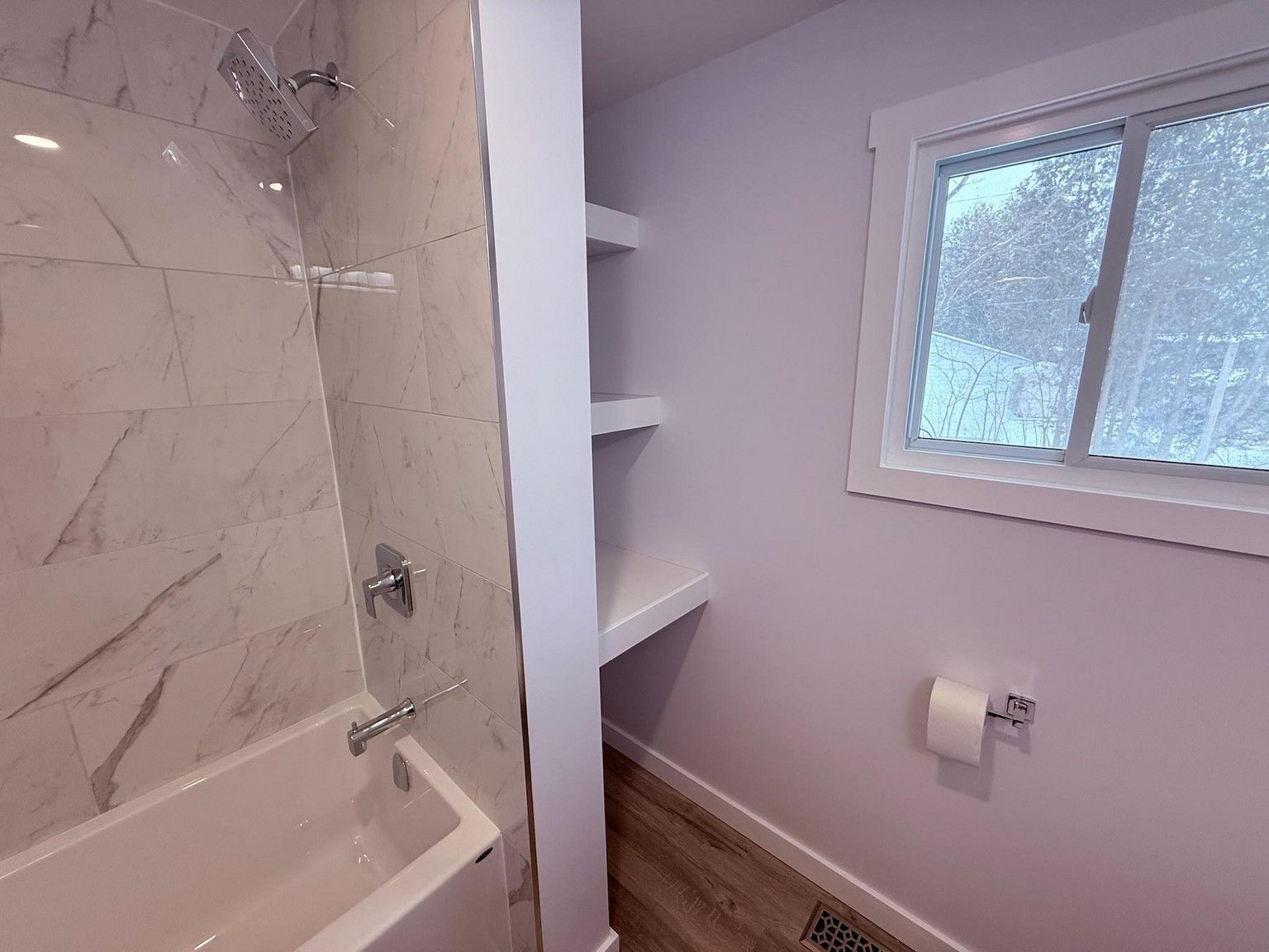 A bathroom with a white marble-tiled bathtub, built-in shelving, a window, and light lavender-colored walls.