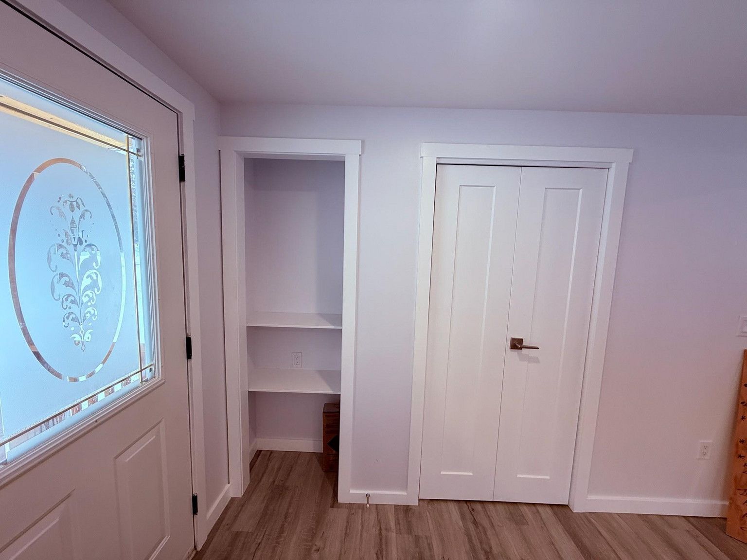 An entryway with a white door, a recessed open shelving unit, and a white double-door closet on a light wood floor.