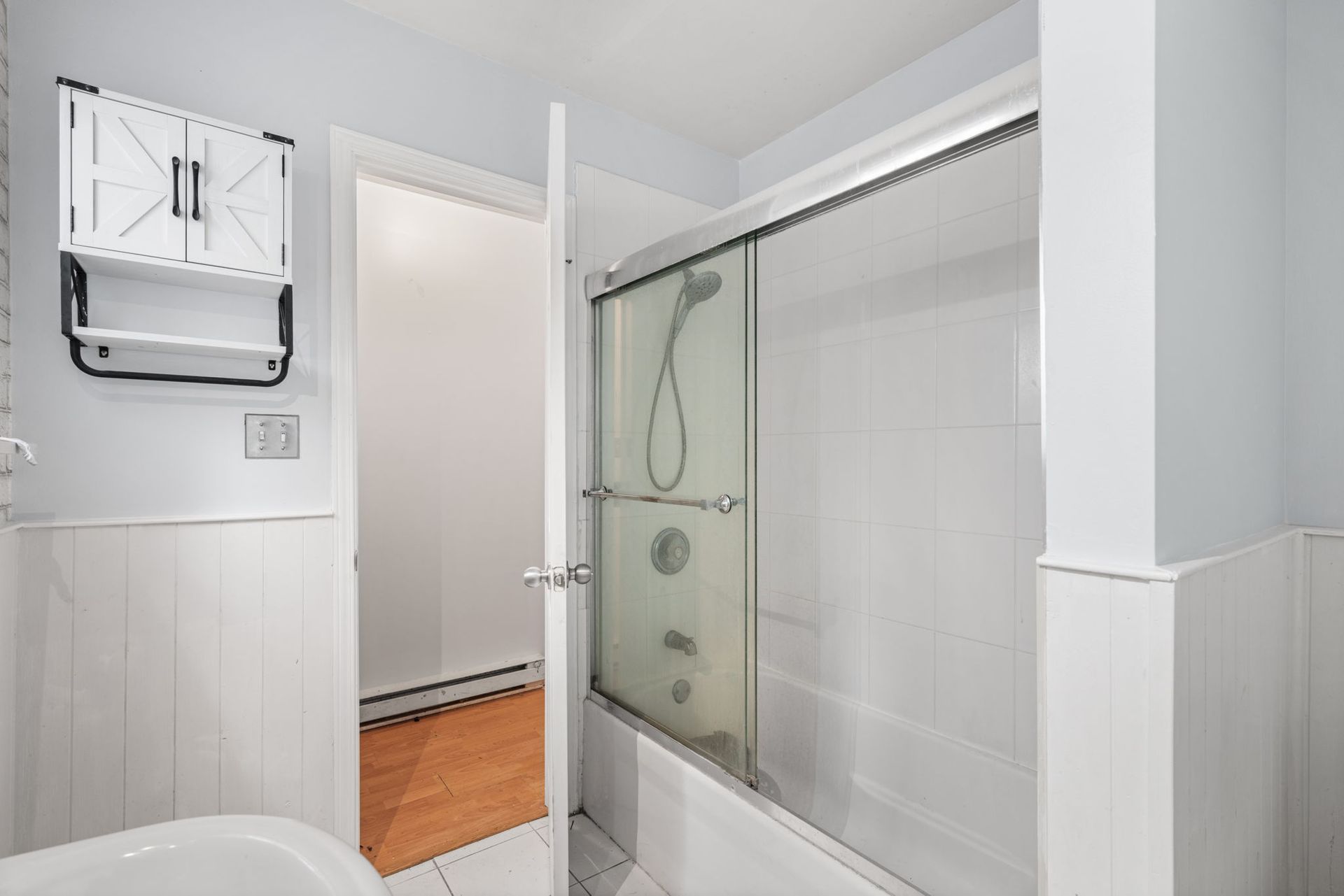 Bathroom with shower, open door, white paneling, and a cabinet.