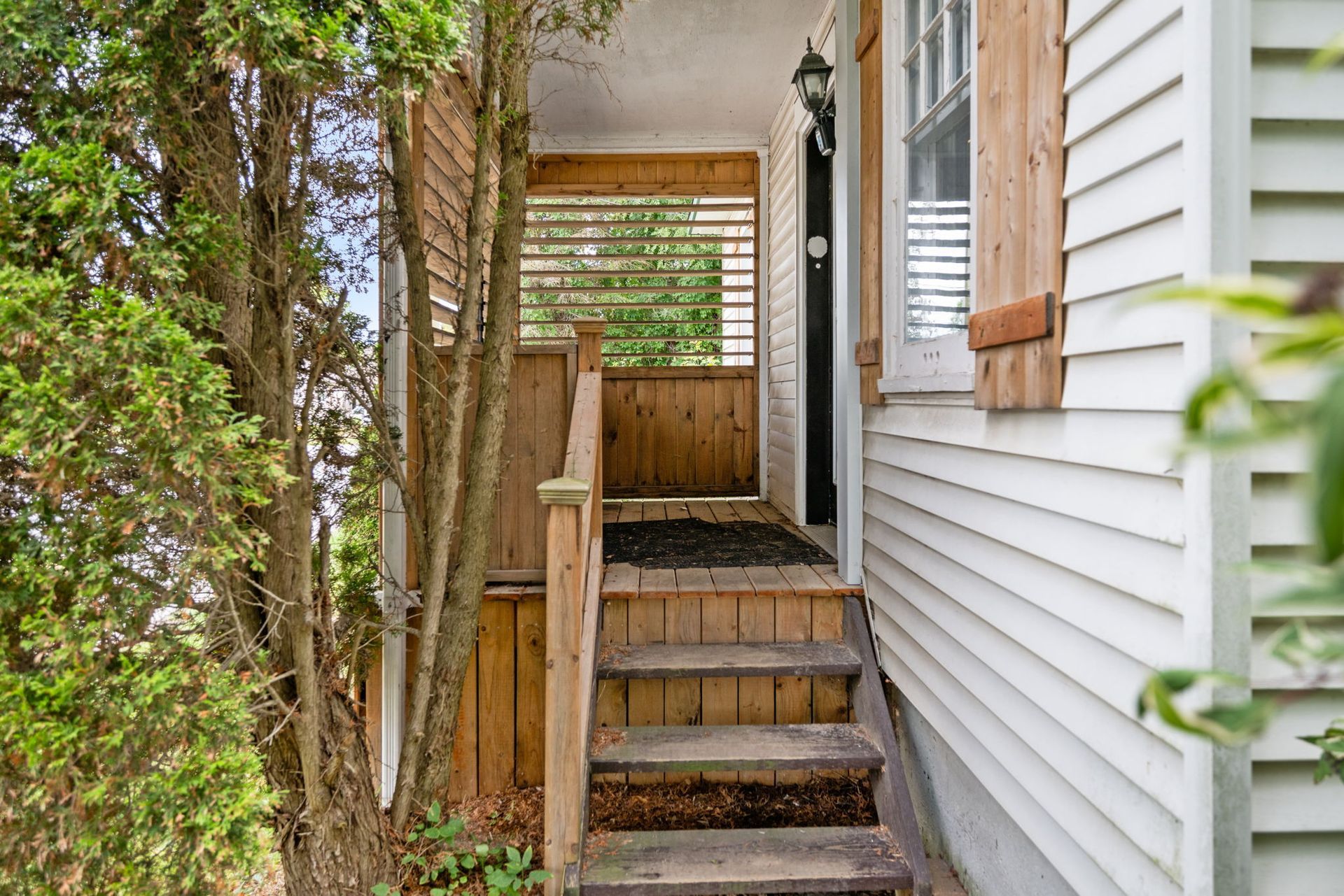 Wooden porch with steps leading to a covered entrance; white siding and green foliage.
