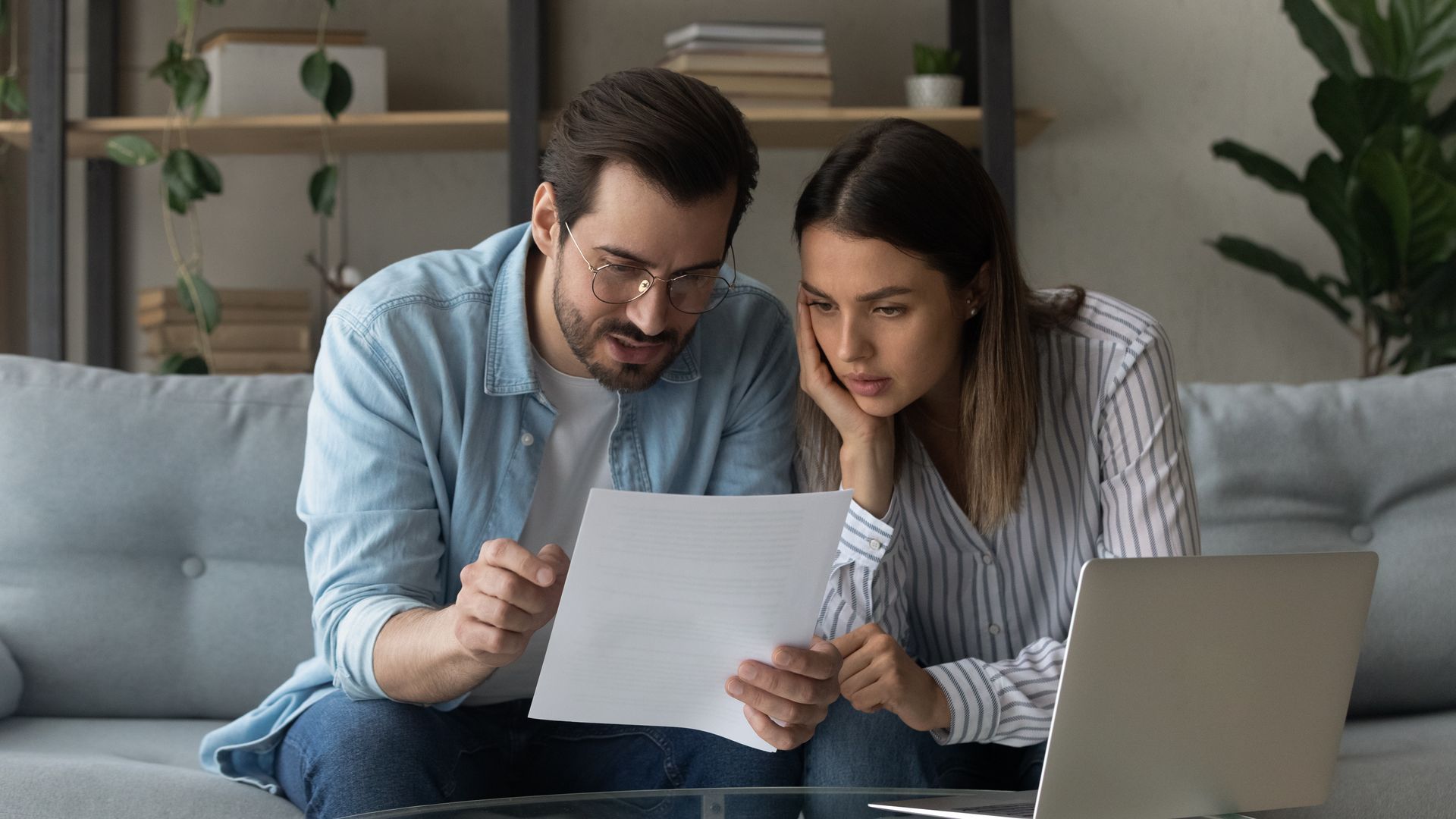 Couple looking at a document, sitting on a couch near a laptop.