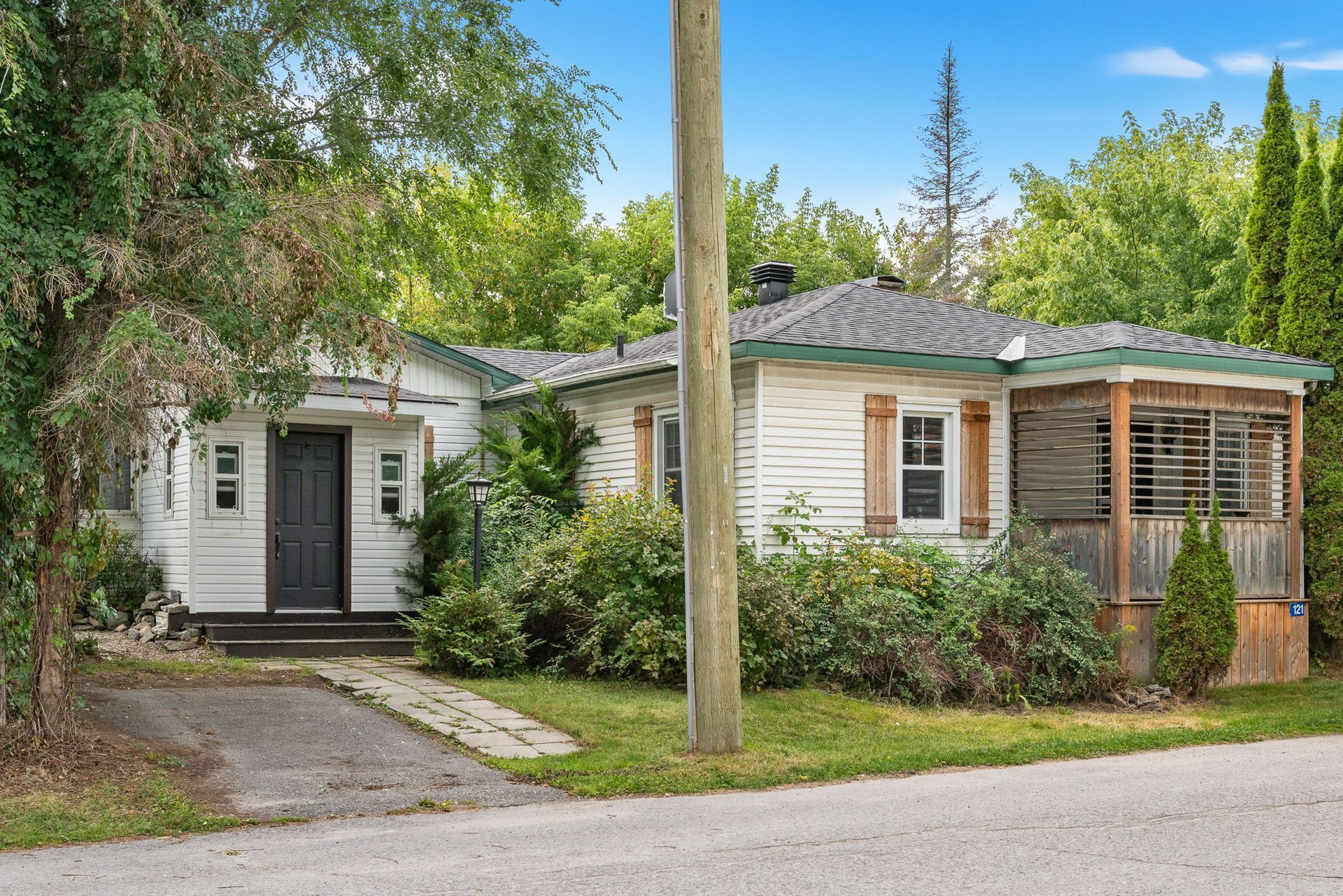 White cottage with black door and screened porch, surrounded by green trees and shrubs, near a paved driveway.