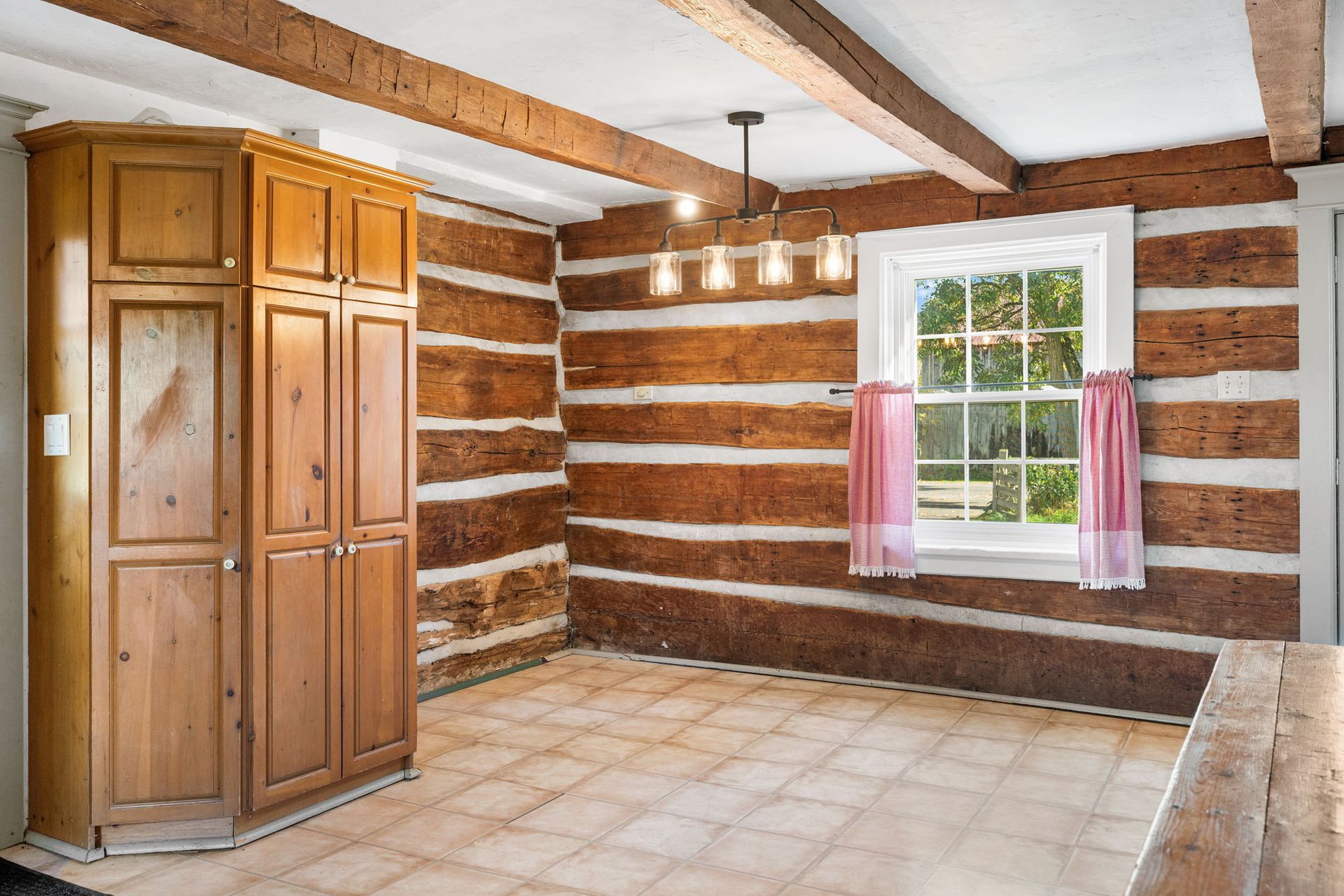 Kitchen with log walls, wooden beams, cabinets, and a window with pink curtains.