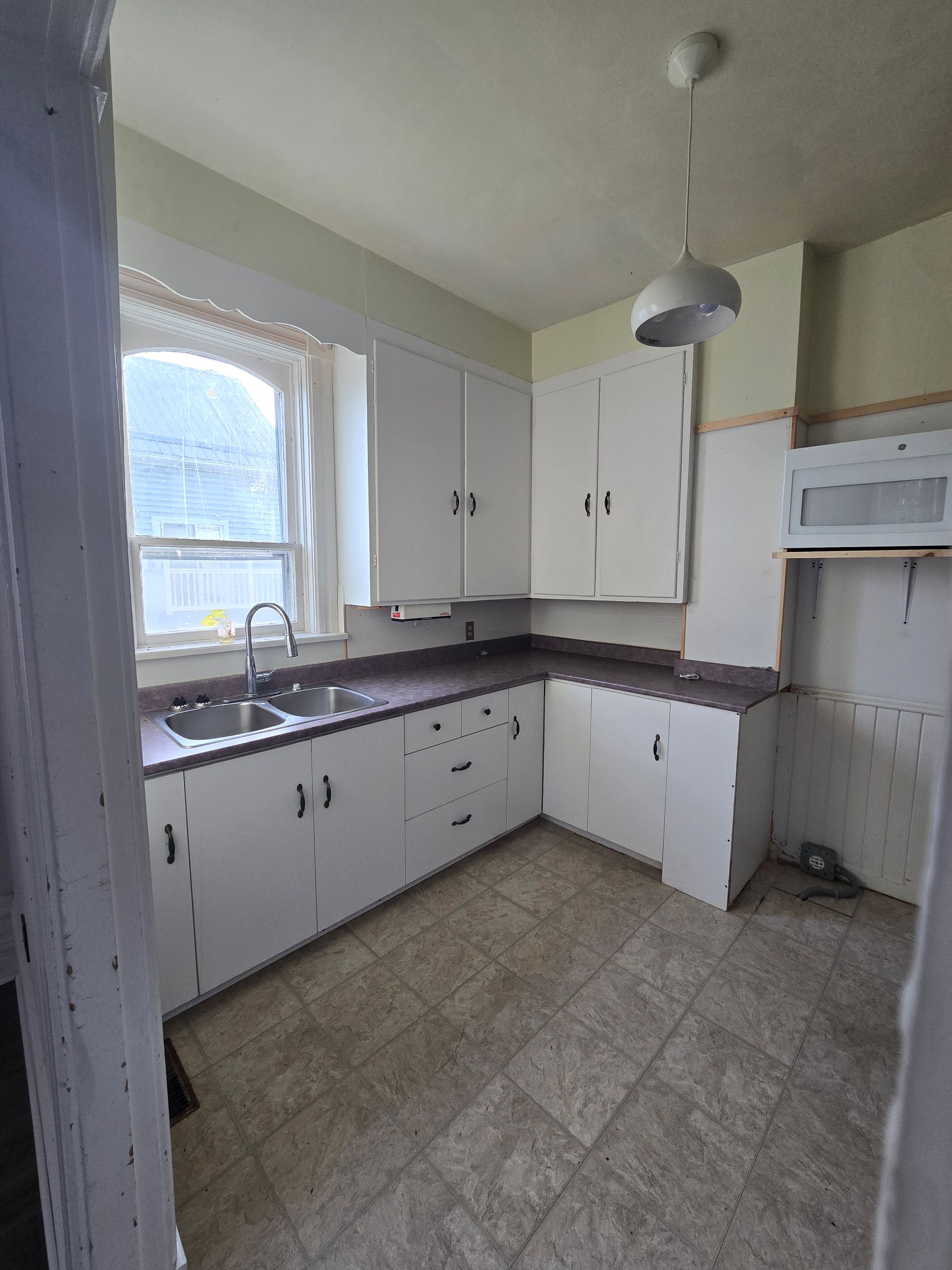 An empty kitchen with white cabinets , a sink , and a window.