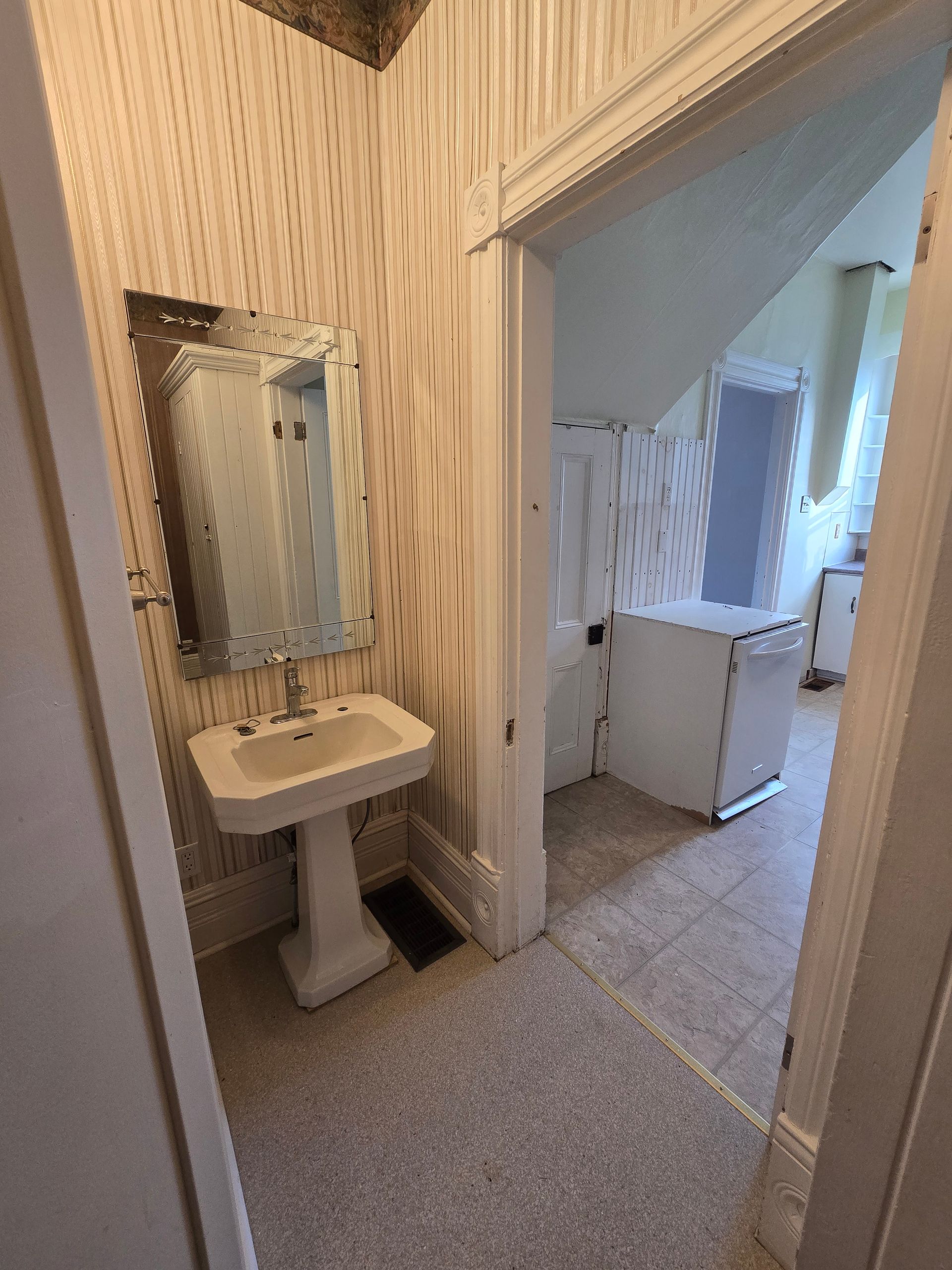 Small bathroom with pedestal sink and mirror, leading to a hallway with a white appliance.