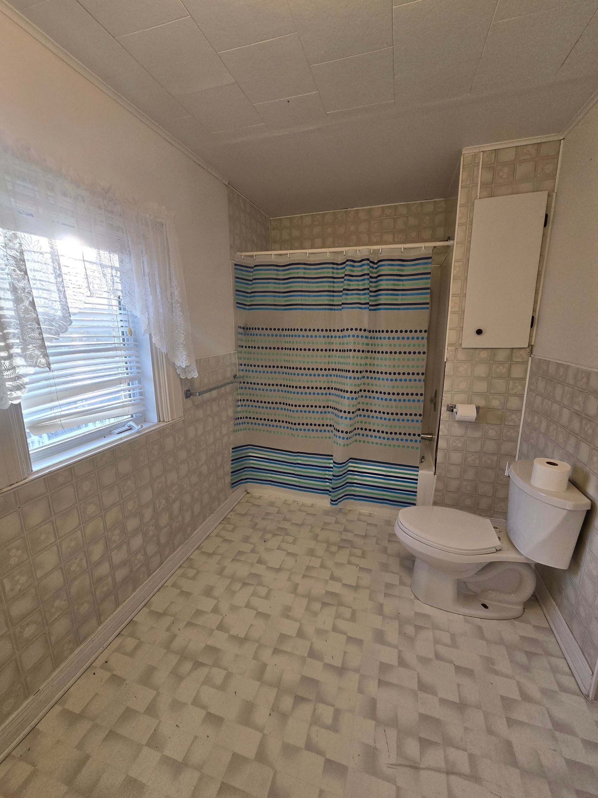 Bathroom with a toilet, shower, and window, tiled in white and gray. A blue shower curtain hangs.