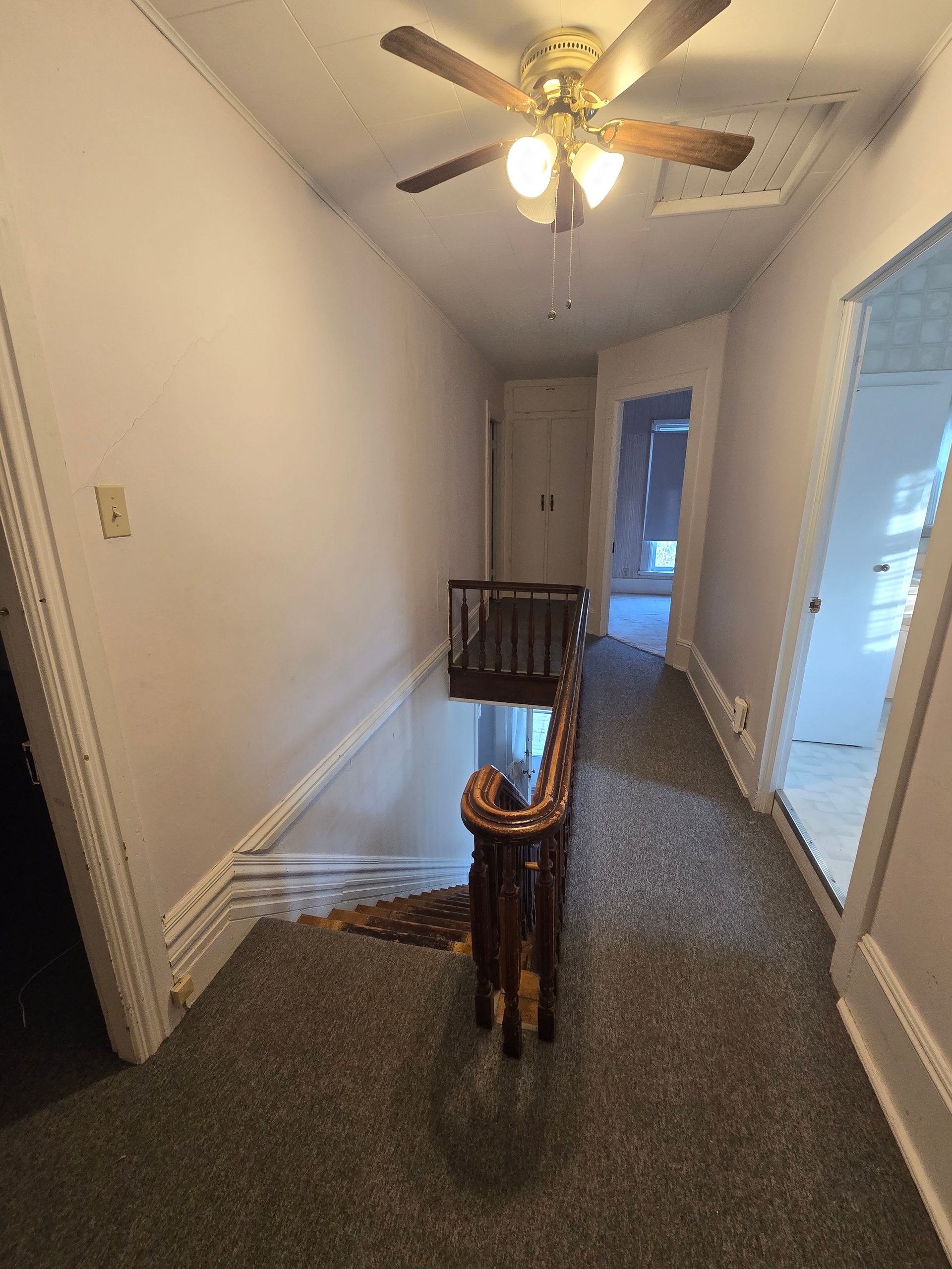 Narrow hallway with a wooden banister, carpeted floor, and doorways. A ceiling fan is visible.