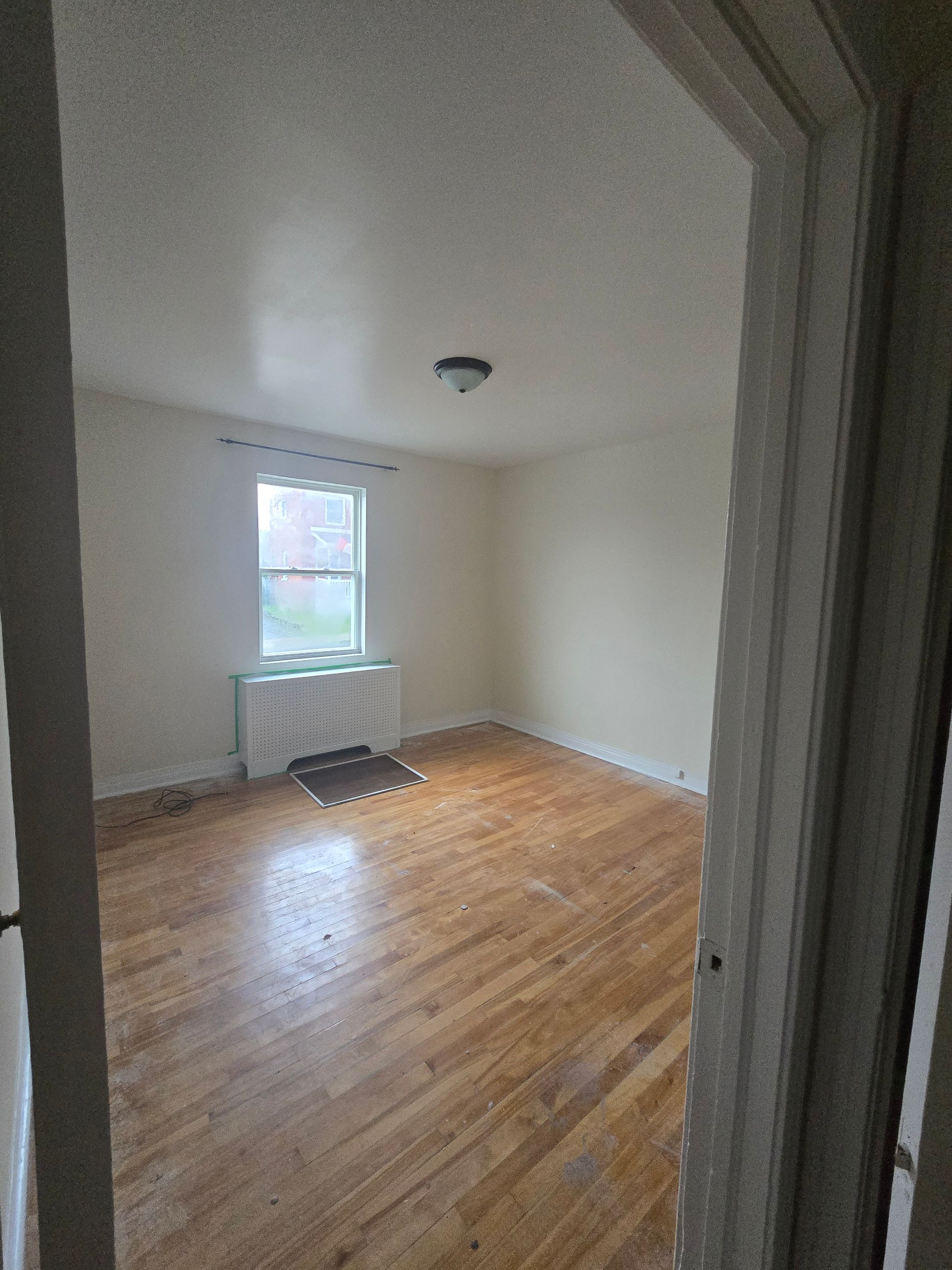 A doorway frames an empty room with wood floors, a window, and a radiator. The walls are painted a light beige.
