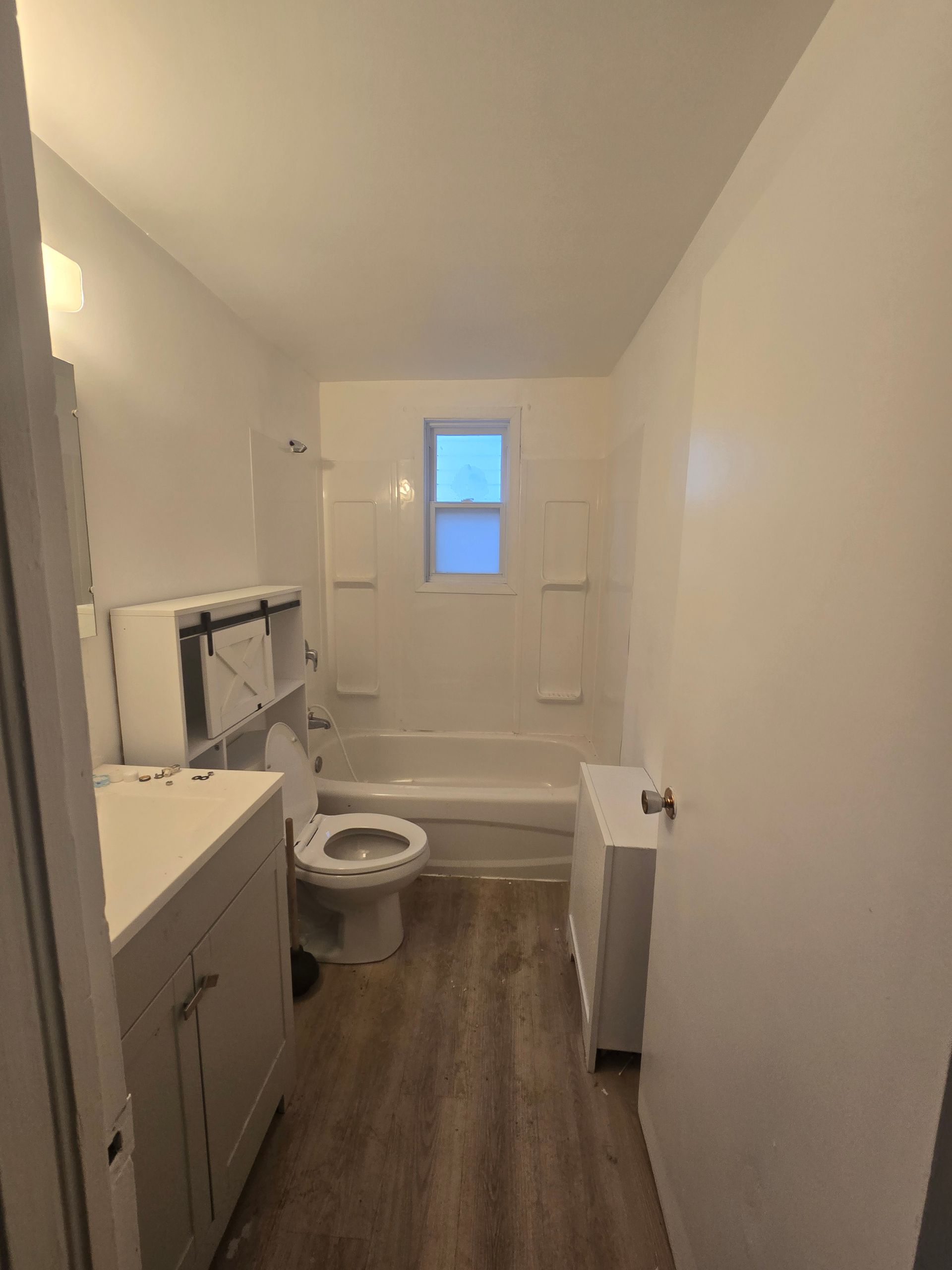 A small, white bathroom with wood-look flooring. Features a toilet, vanity, bathtub/shower, and a small window.
