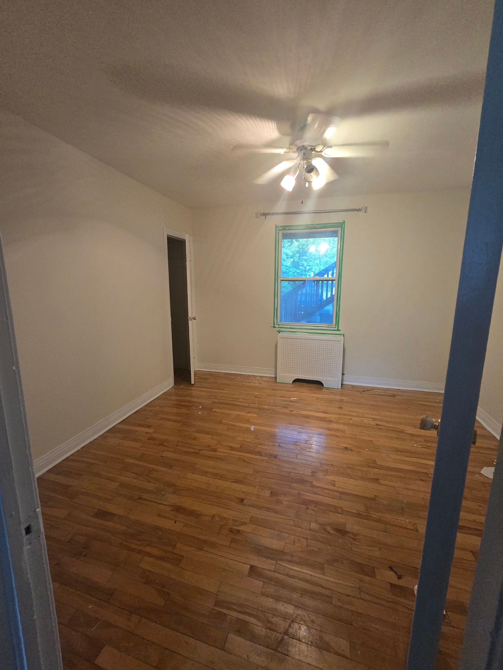 Empty bedroom with hardwood floors, a window, door, and ceiling fan. The walls are beige and the room is dimly lit.