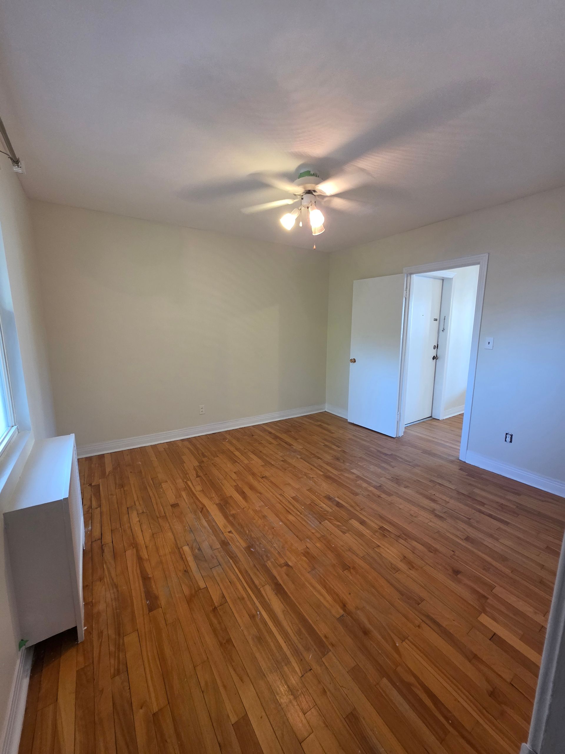 Empty room with hardwood floors, white walls, and a ceiling fan. A white cabinet sits near a window.