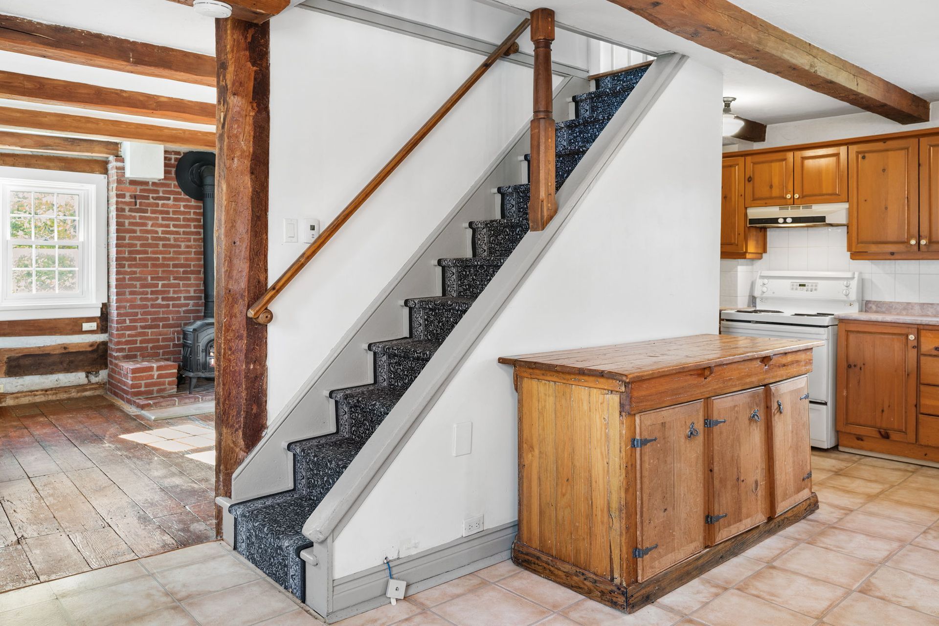 Staircase with carpeted steps, wooden handrail, leading to the upper floor, next to a wood cabinet in a kitchen.