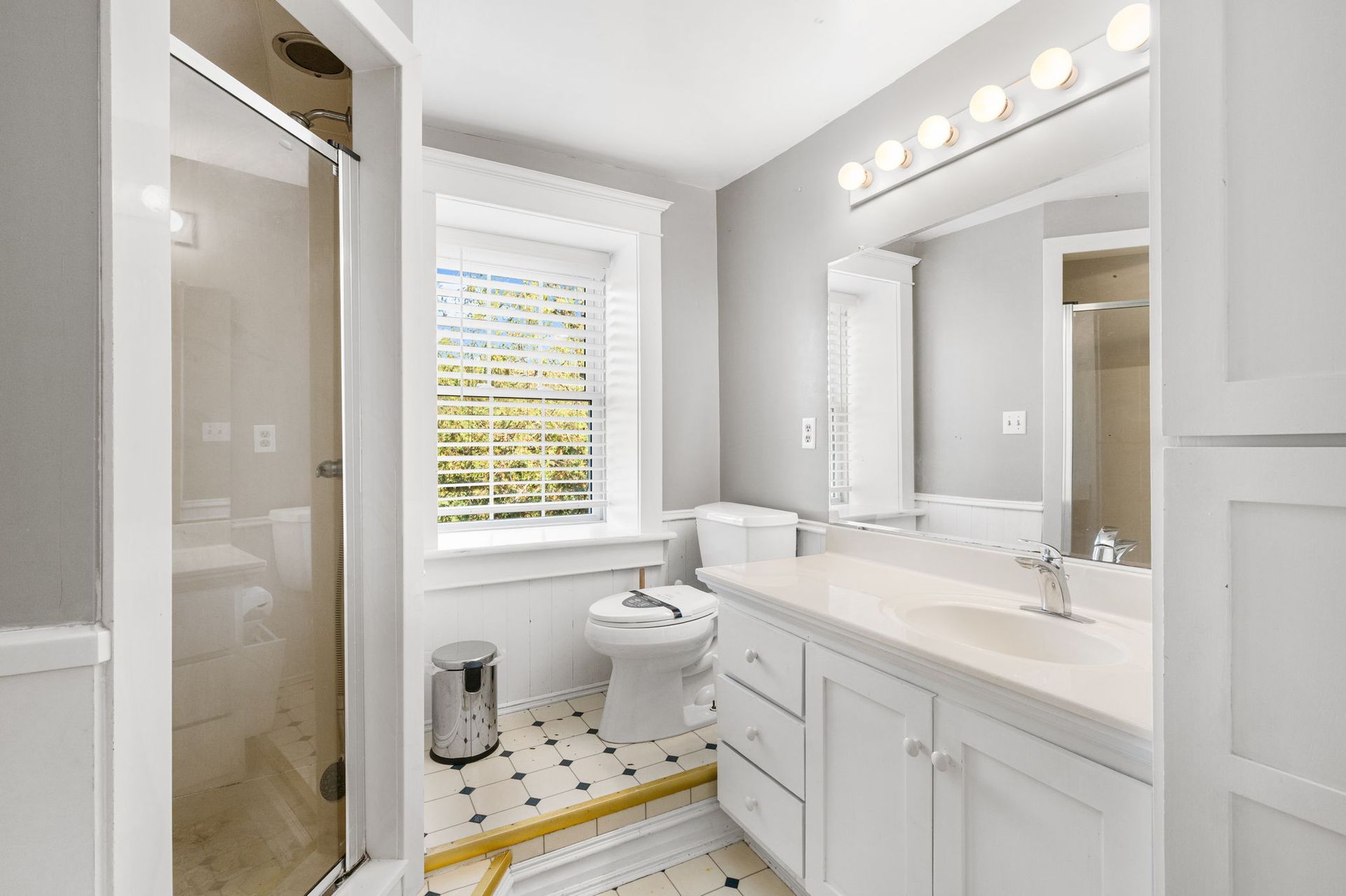 Bathroom with white cabinets, toilet, and shower. Black and white tiled floor. Window with blinds.