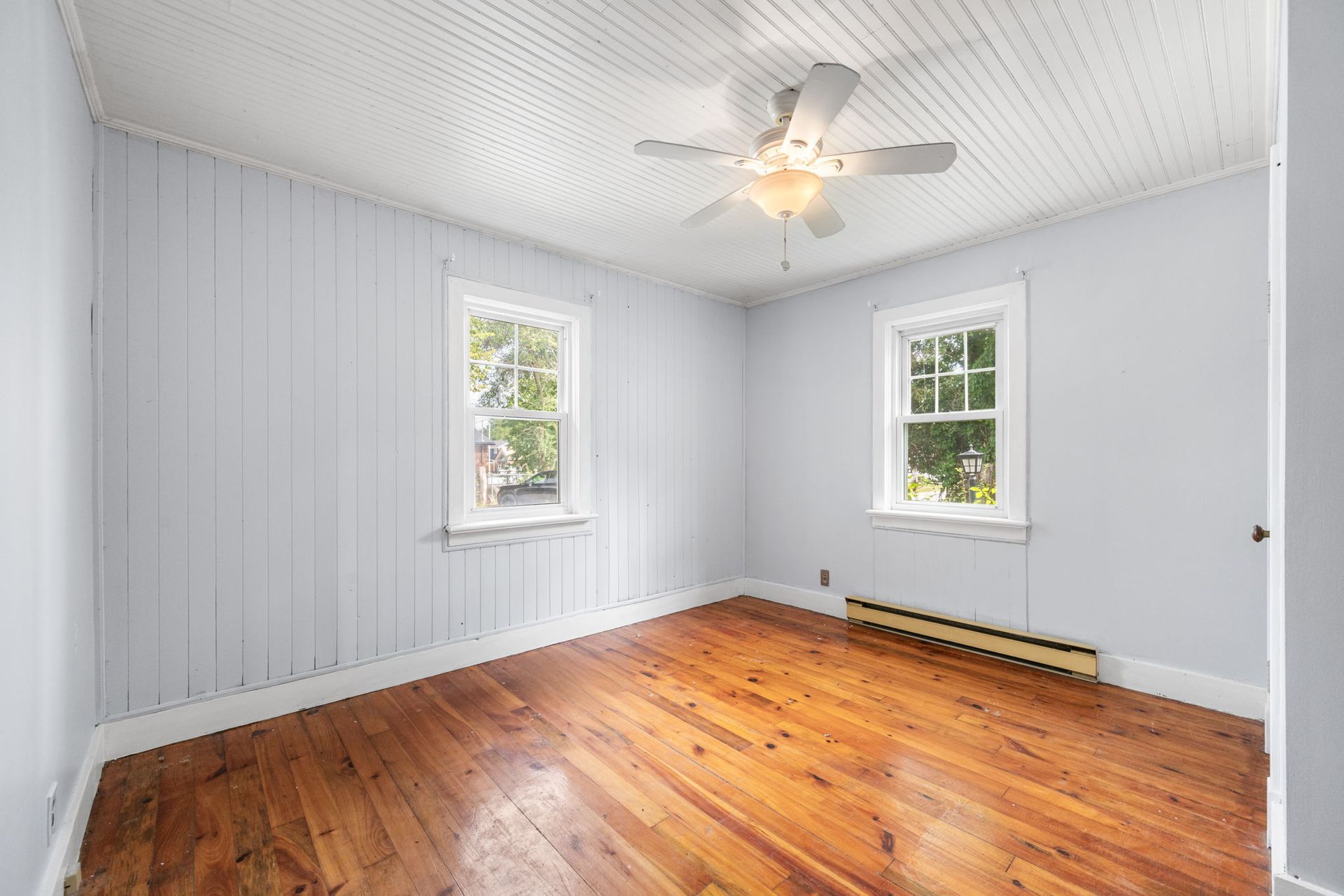 Empty room with hardwood floors, light blue walls, white trim, and a ceiling fan. Two windows.