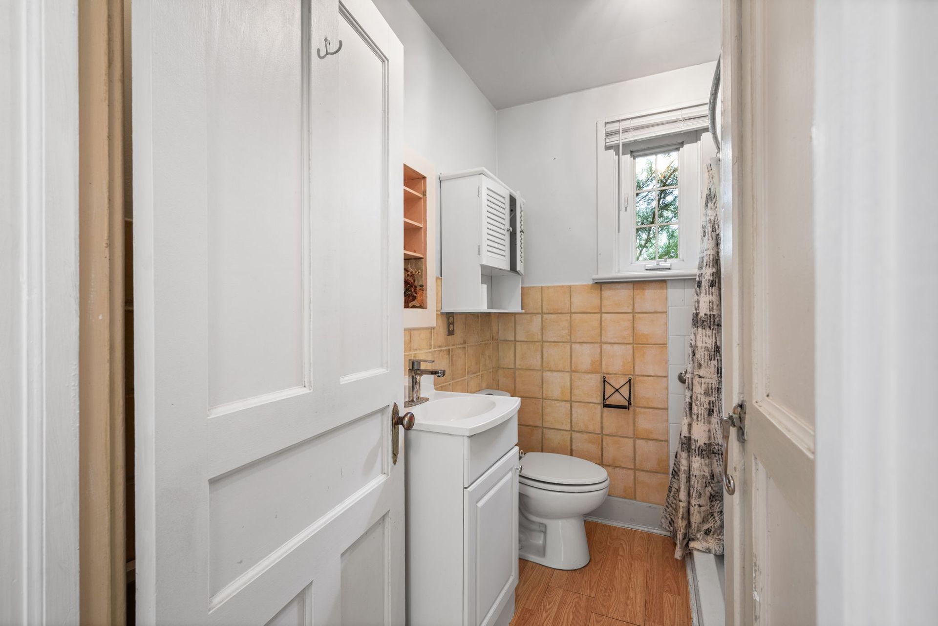 Small, white bathroom with a sink, toilet, and tiled wall.  A window is on the right side.