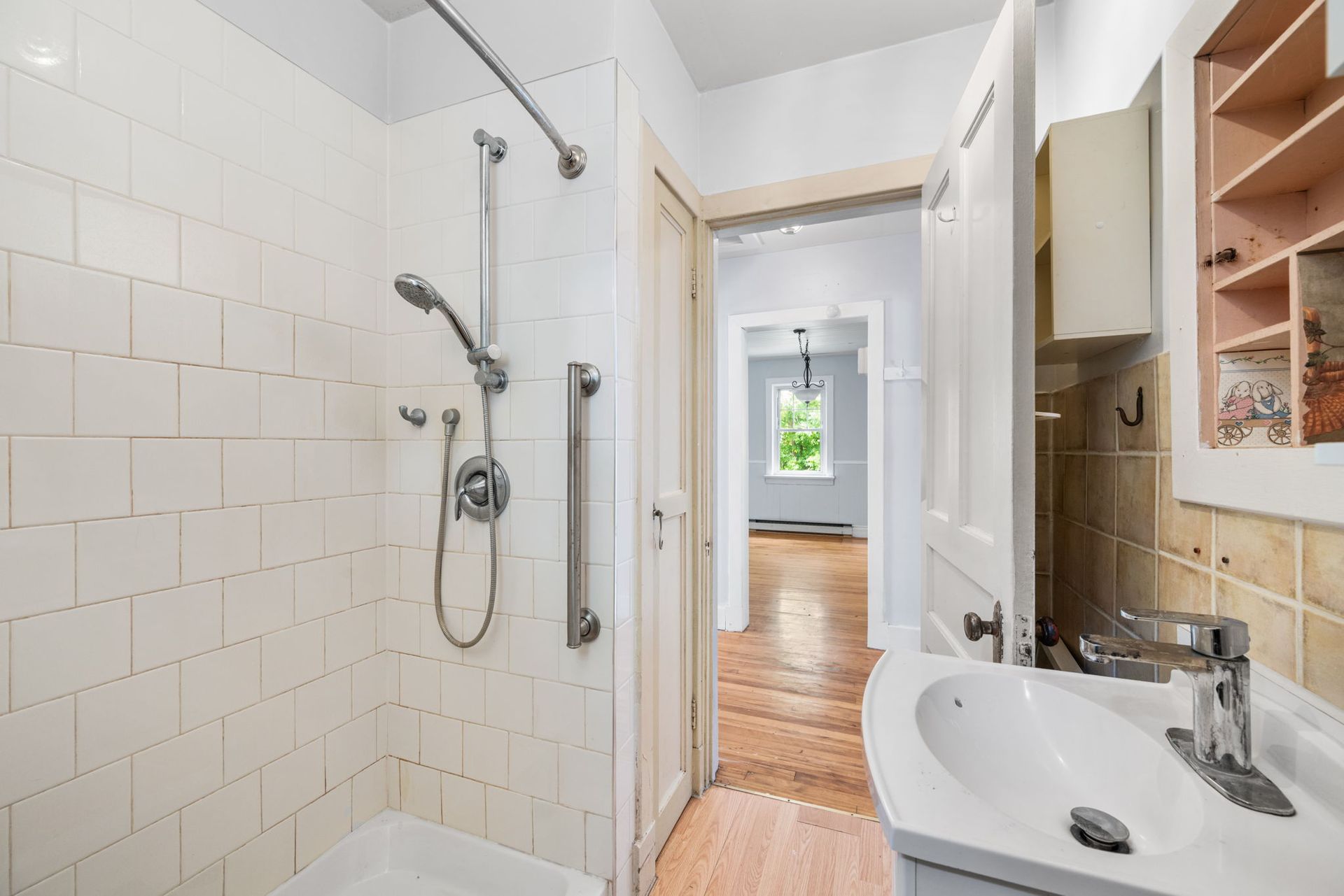 Bathroom with white tiled shower, sink, and doorway leading to a room with wood floor.