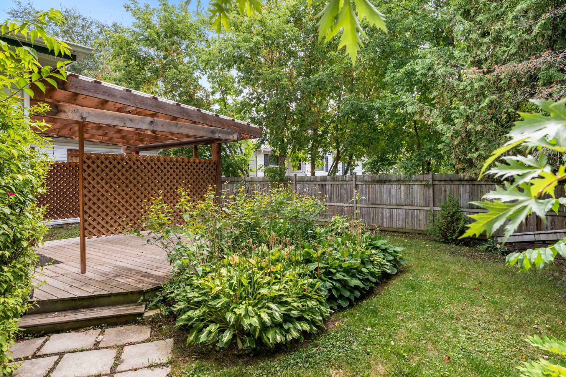 Backyard with wooden deck, trellis, and lush greenery. A wooden fence borders the yard.