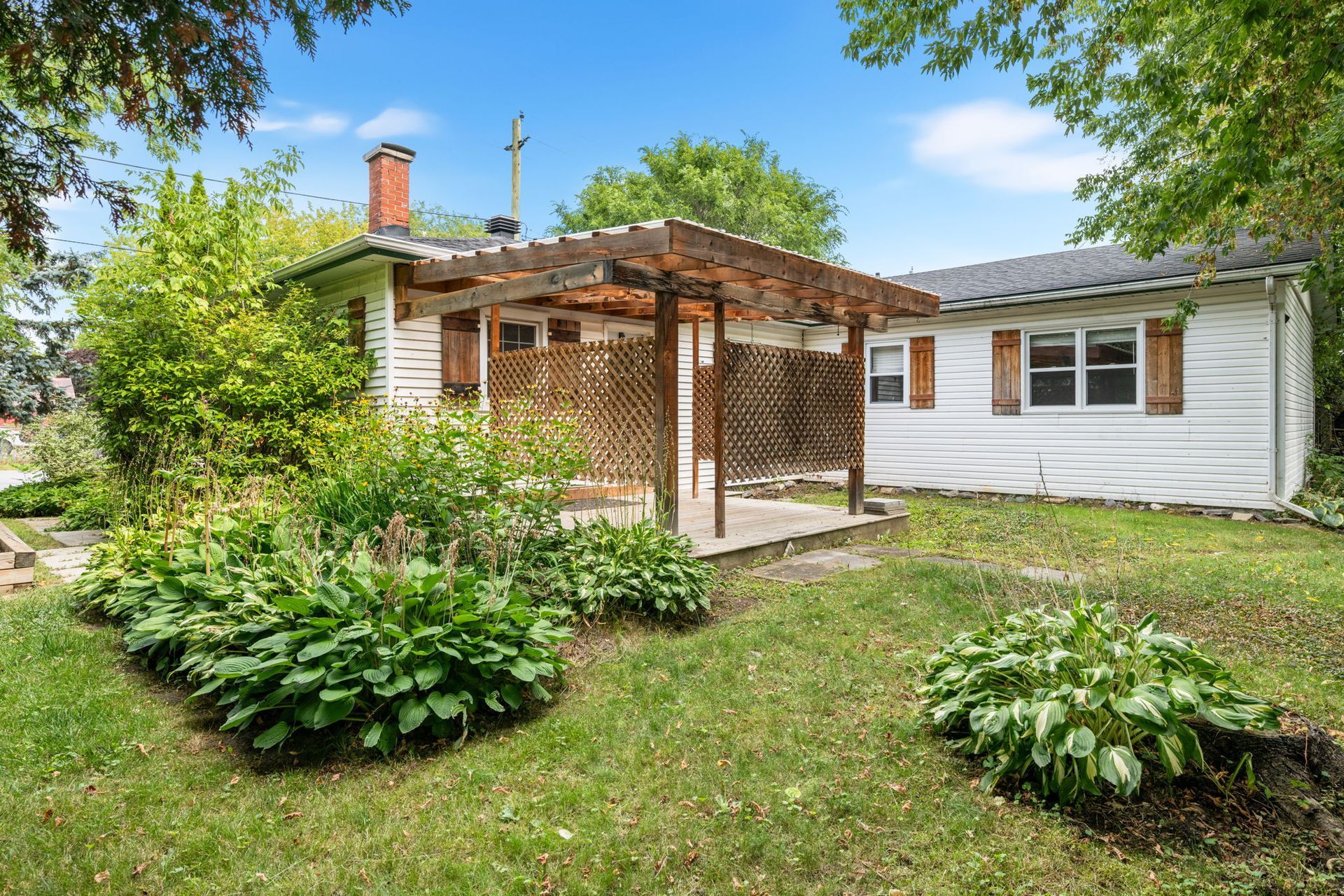 Small white house with wooden porch and garden, surrounded by greenery and a grassy yard.
