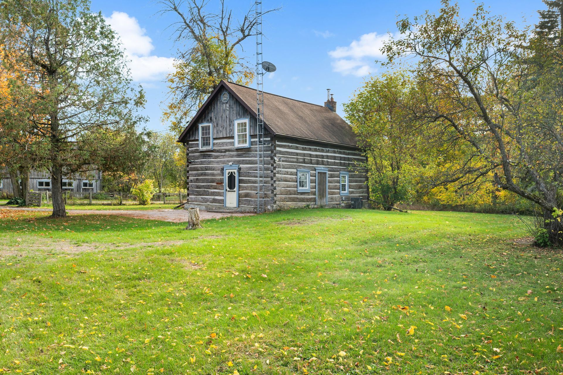 Log cabin house in a grassy yard, surrounded by trees with autumn foliage under a blue sky.