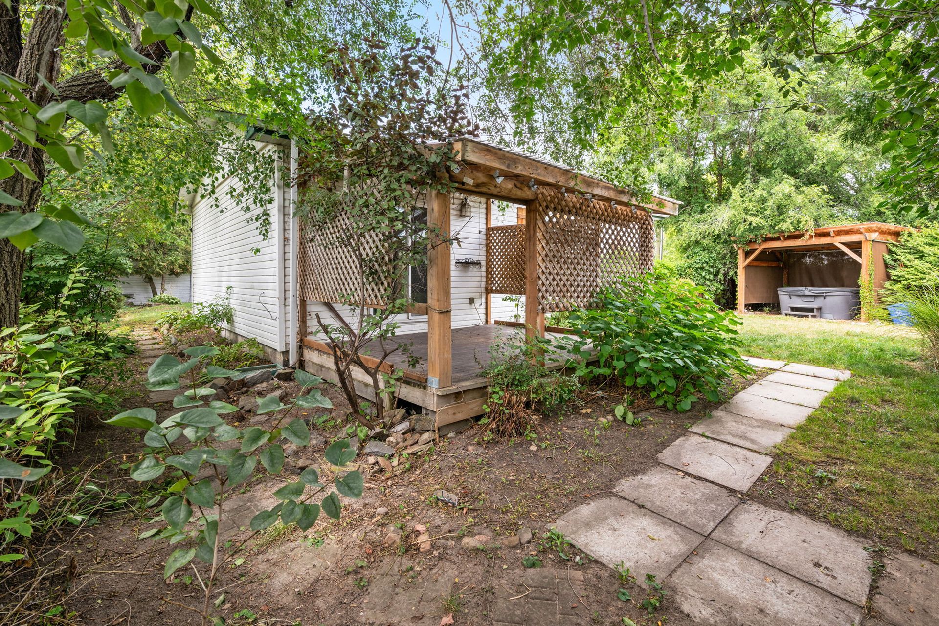 Backyard with small wooden porch, path, and hot tub under a pergola. Lush greenery surrounds the structures.