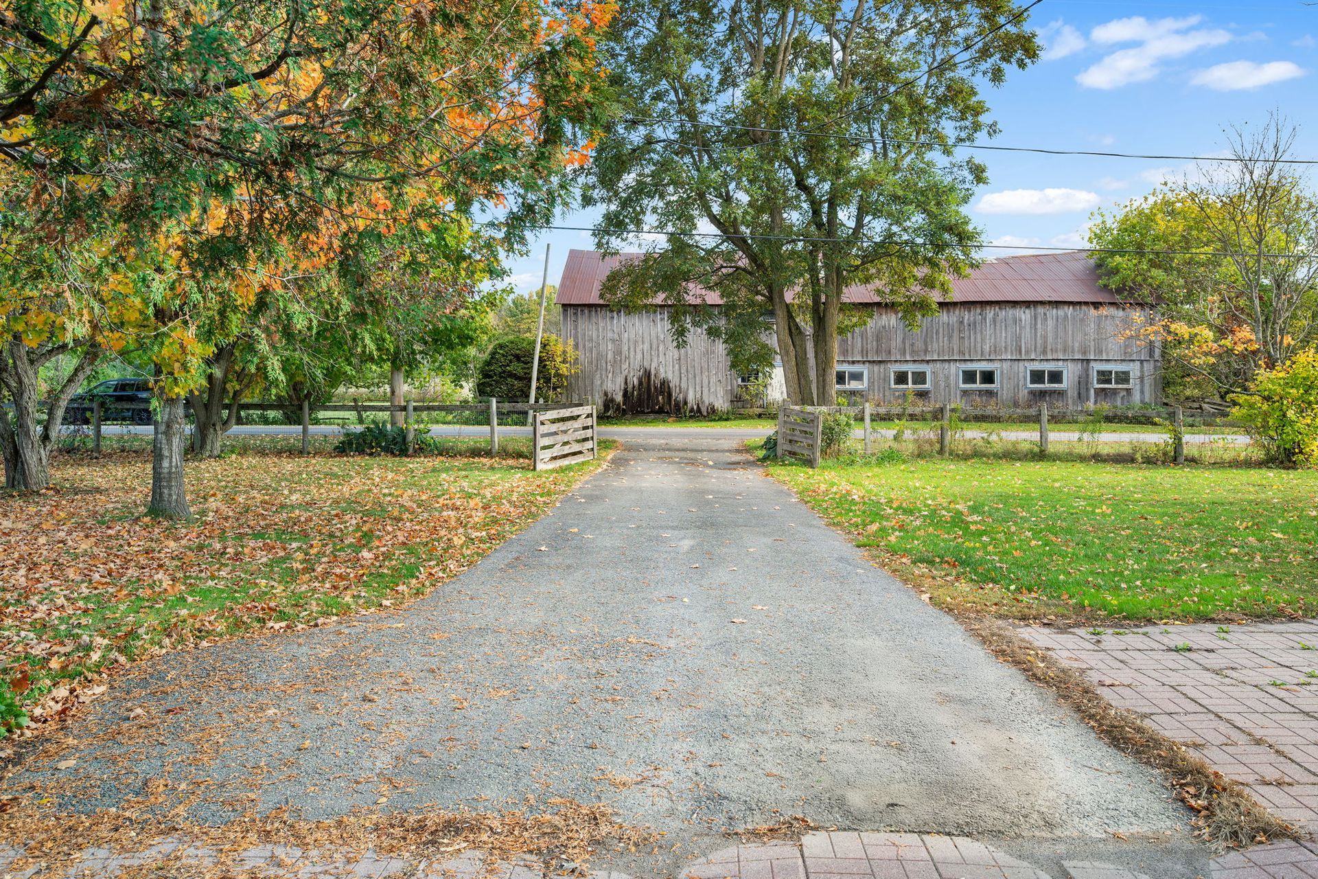 Gravel driveway leading to a weathered wooden barn, framed by trees with fall foliage under a blue sky.