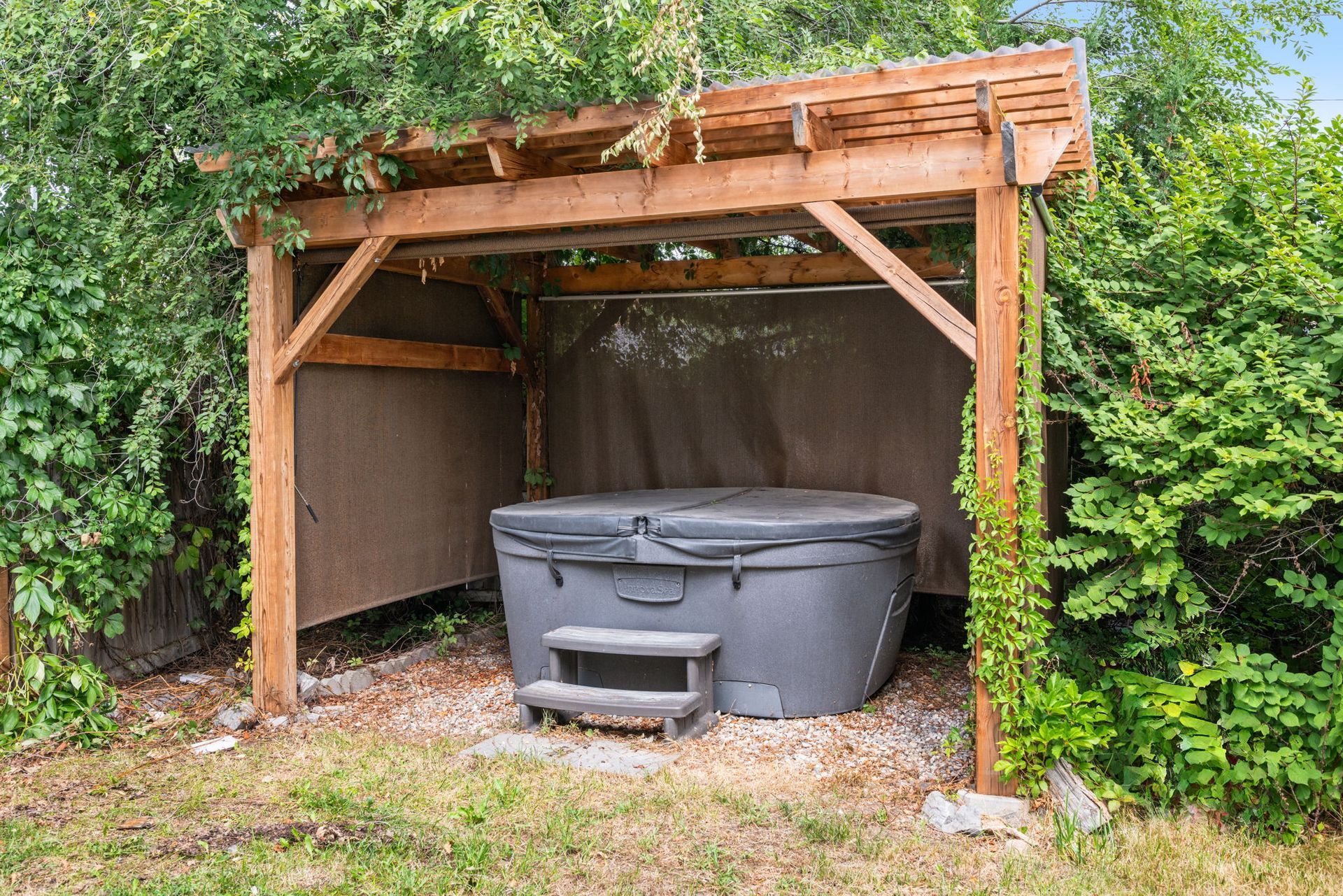 Hot tub under a wooden pergola in a backyard, surrounded by greenery.