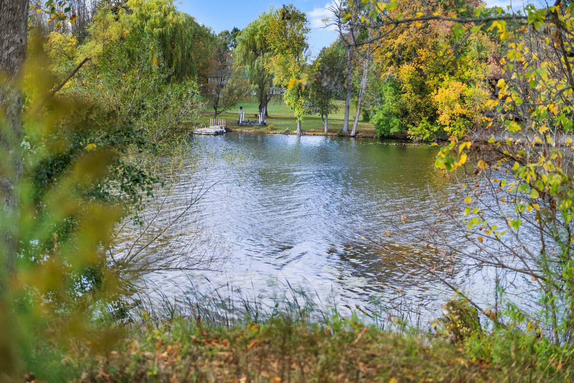 A lake surrounded by trees with fall foliage. People stand on a small pier in the distance.