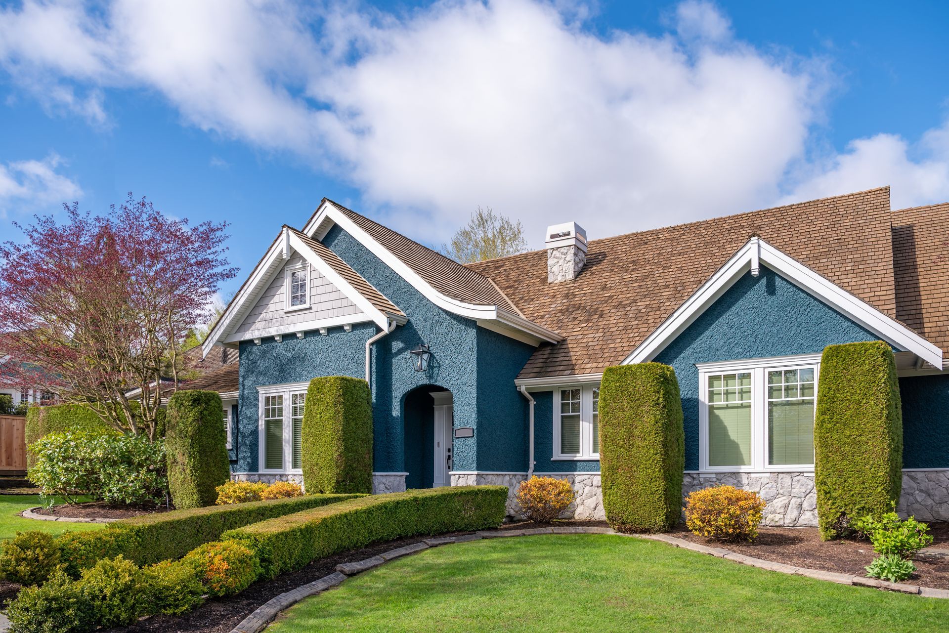 Blue suburban house with white trim, manicured lawn, and bright hedges under a partly cloudy sky