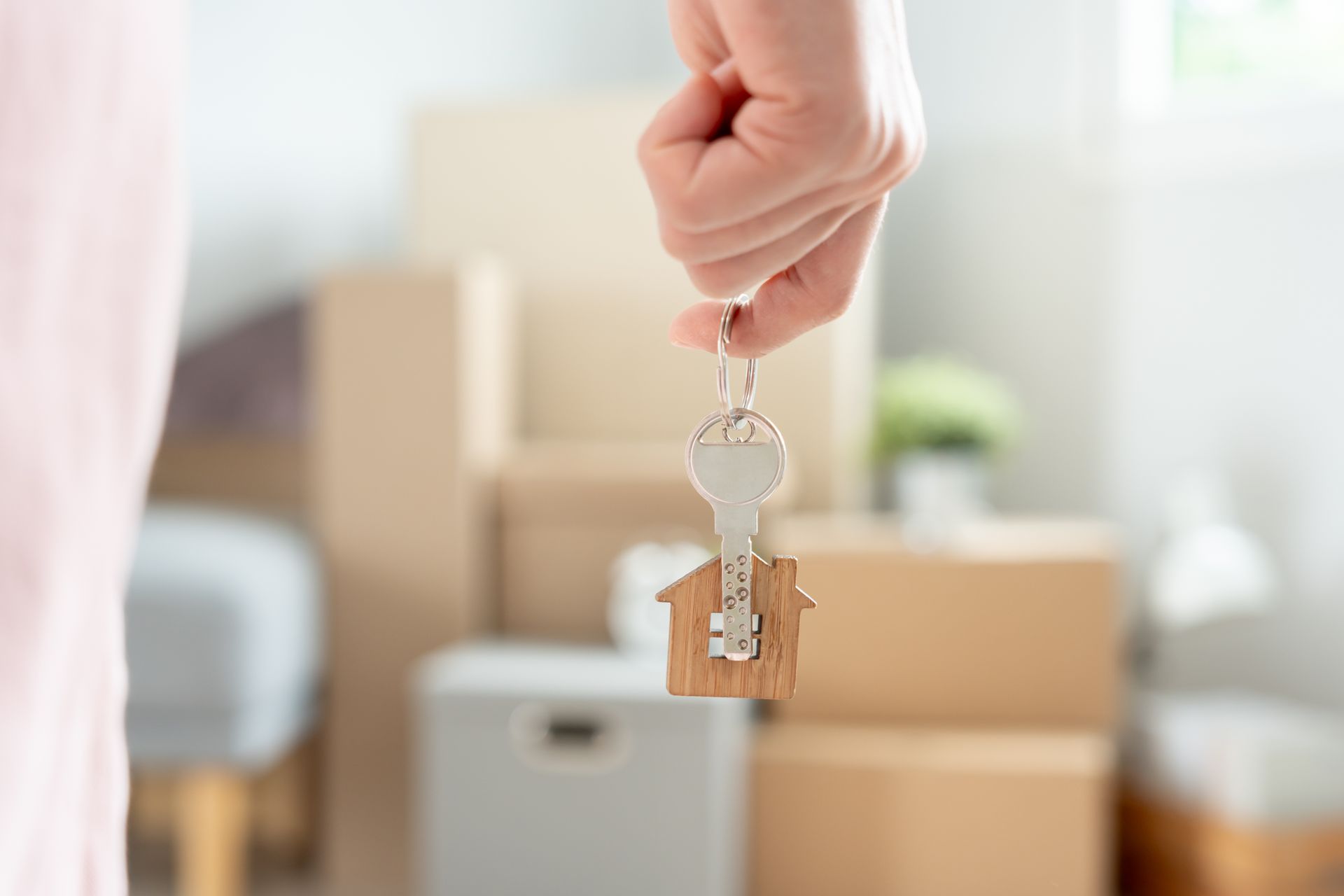 Hand holding house key with wooden house charm, boxes in background.