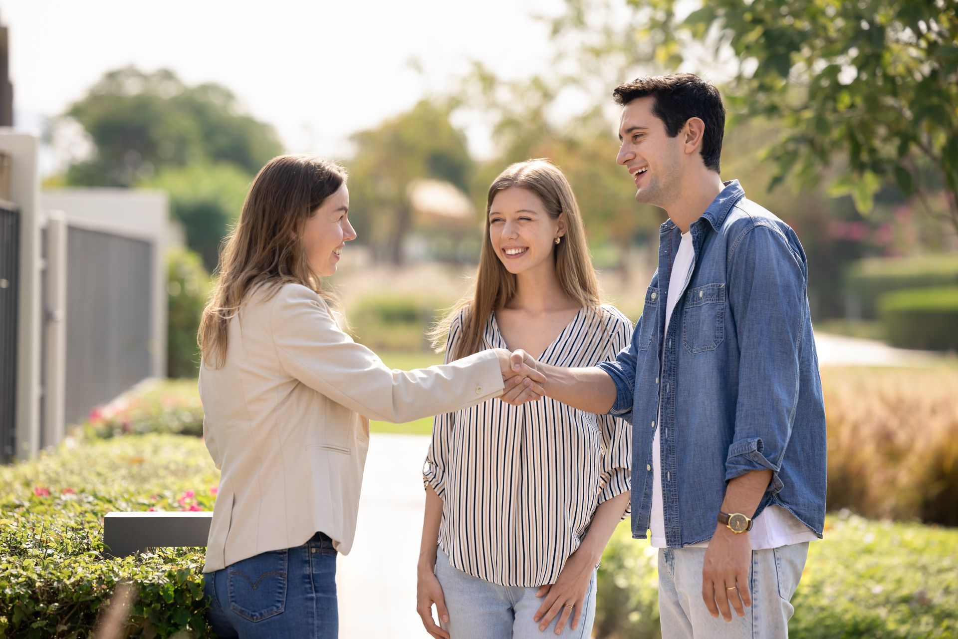Three people smiling and shaking hands outdoors on a sunny campus walkway.