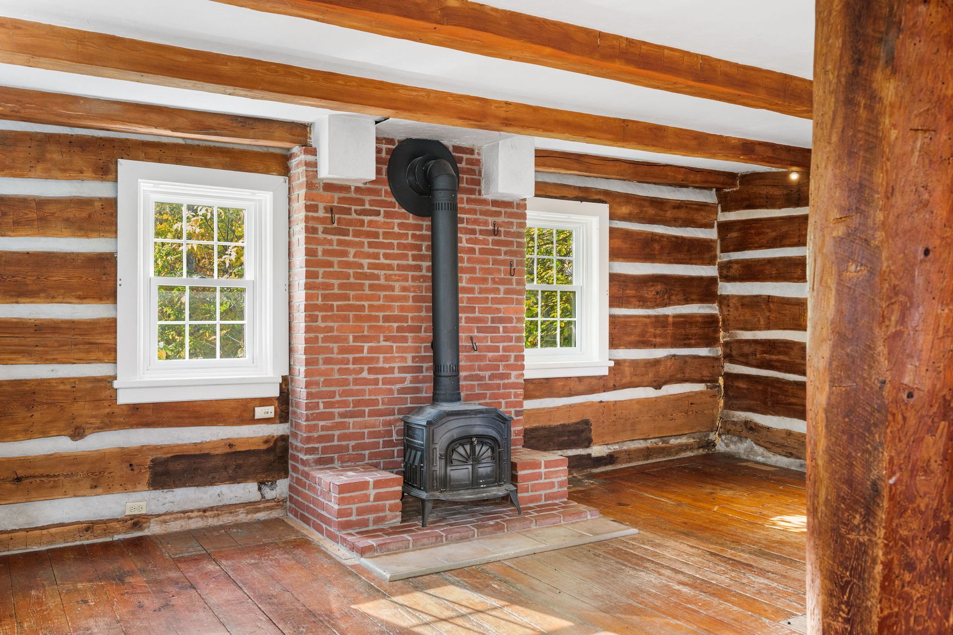 Interior of a log cabin with a brick fireplace and stove, two windows, and wooden beams.