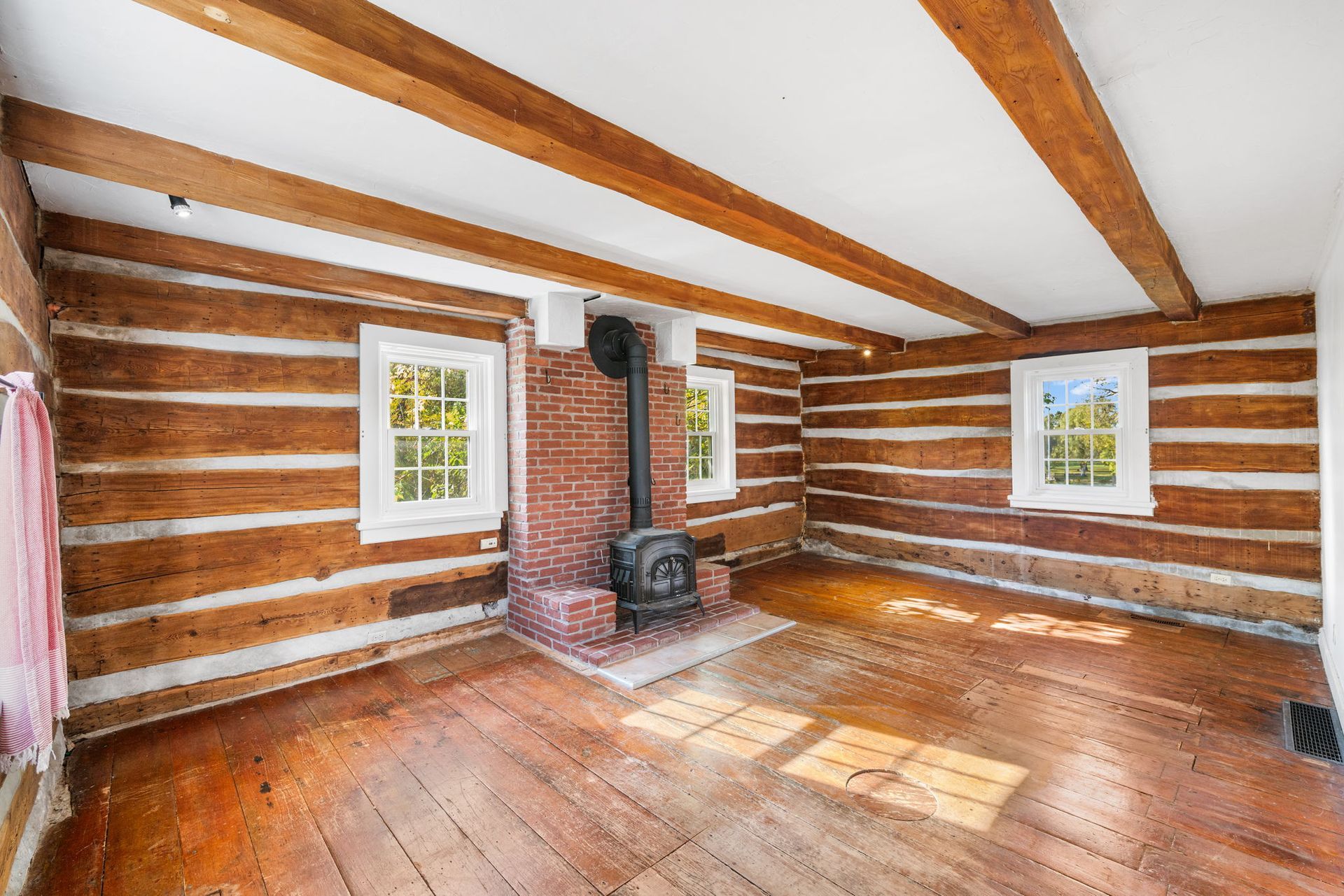 Log cabin interior with exposed wooden beams, brick fireplace, and windows.