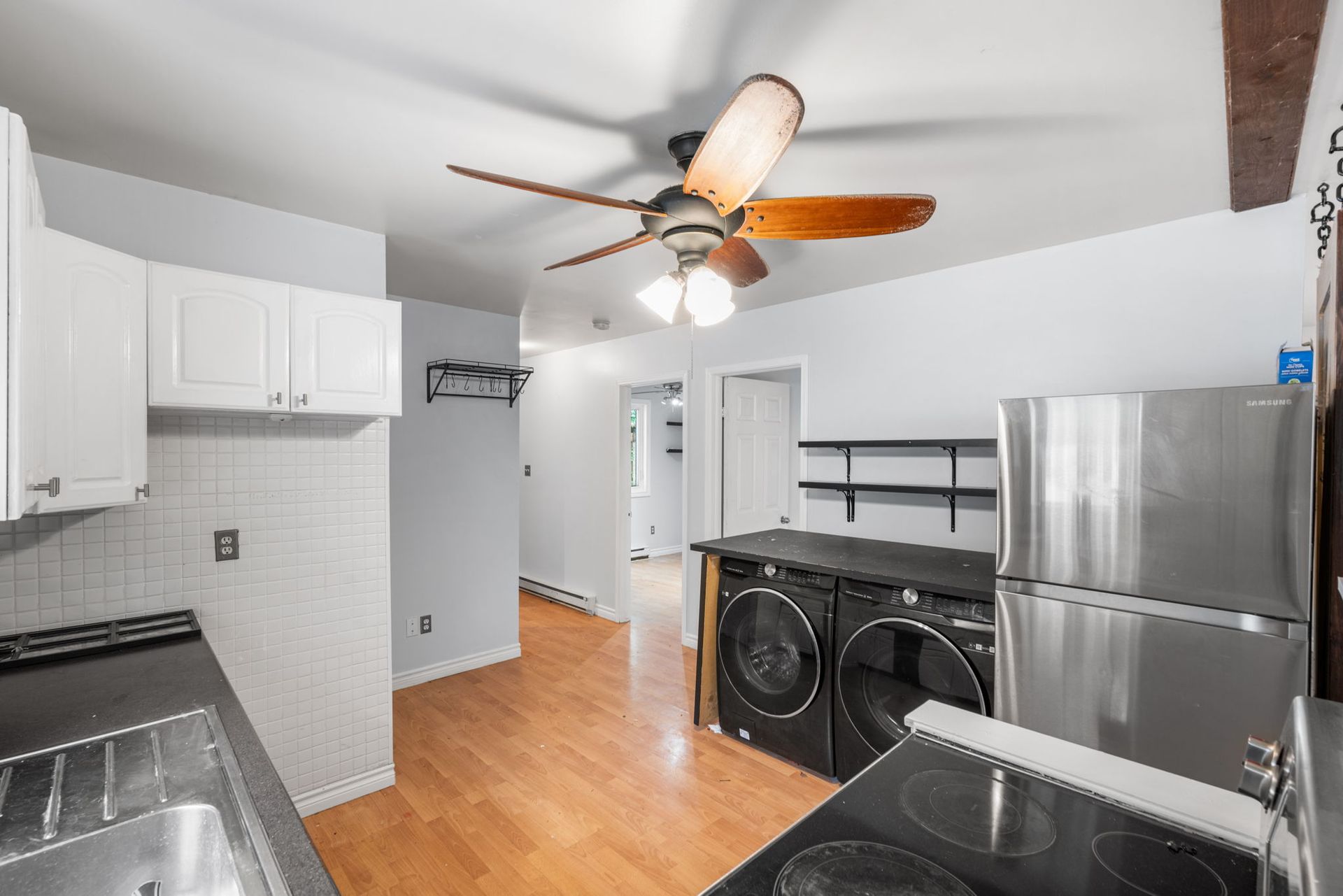 Kitchen with white cabinets, appliances, and a hallway. Wooden floors and a ceiling fan.