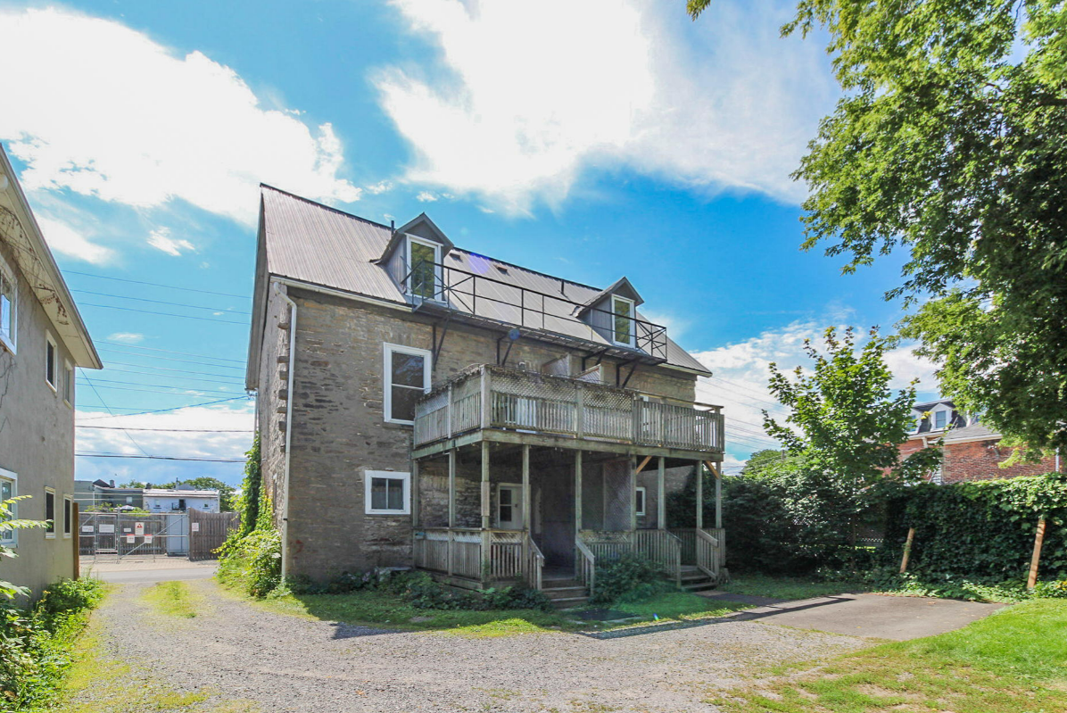 Two-story stone building with balcony, grey roof, and blue sky.